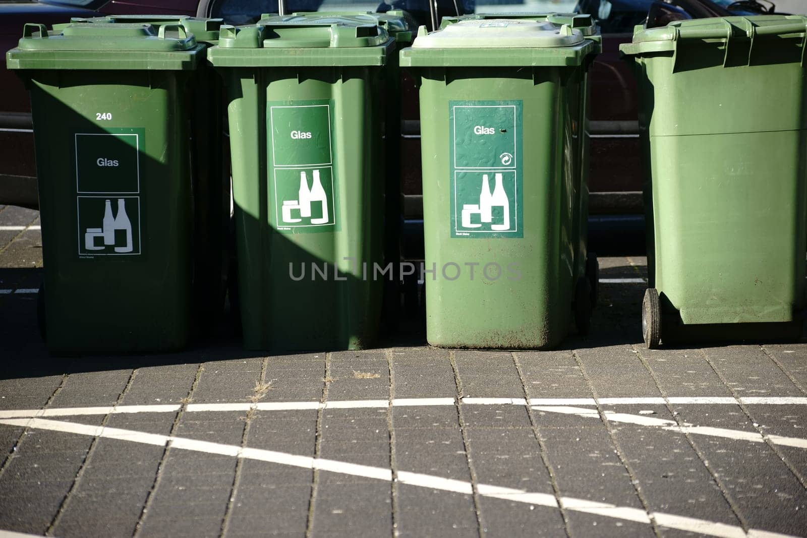 A set of green garbage containers for glass waste stands on a sidewalk in the sun.