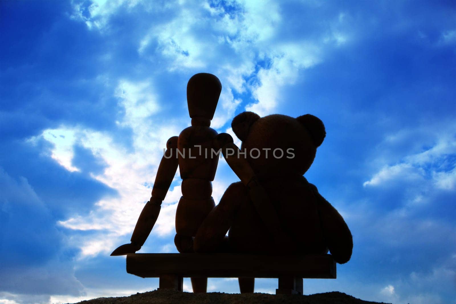 Wooden figure and teddy bear sitting on wooden bench in front of blue sky in back view