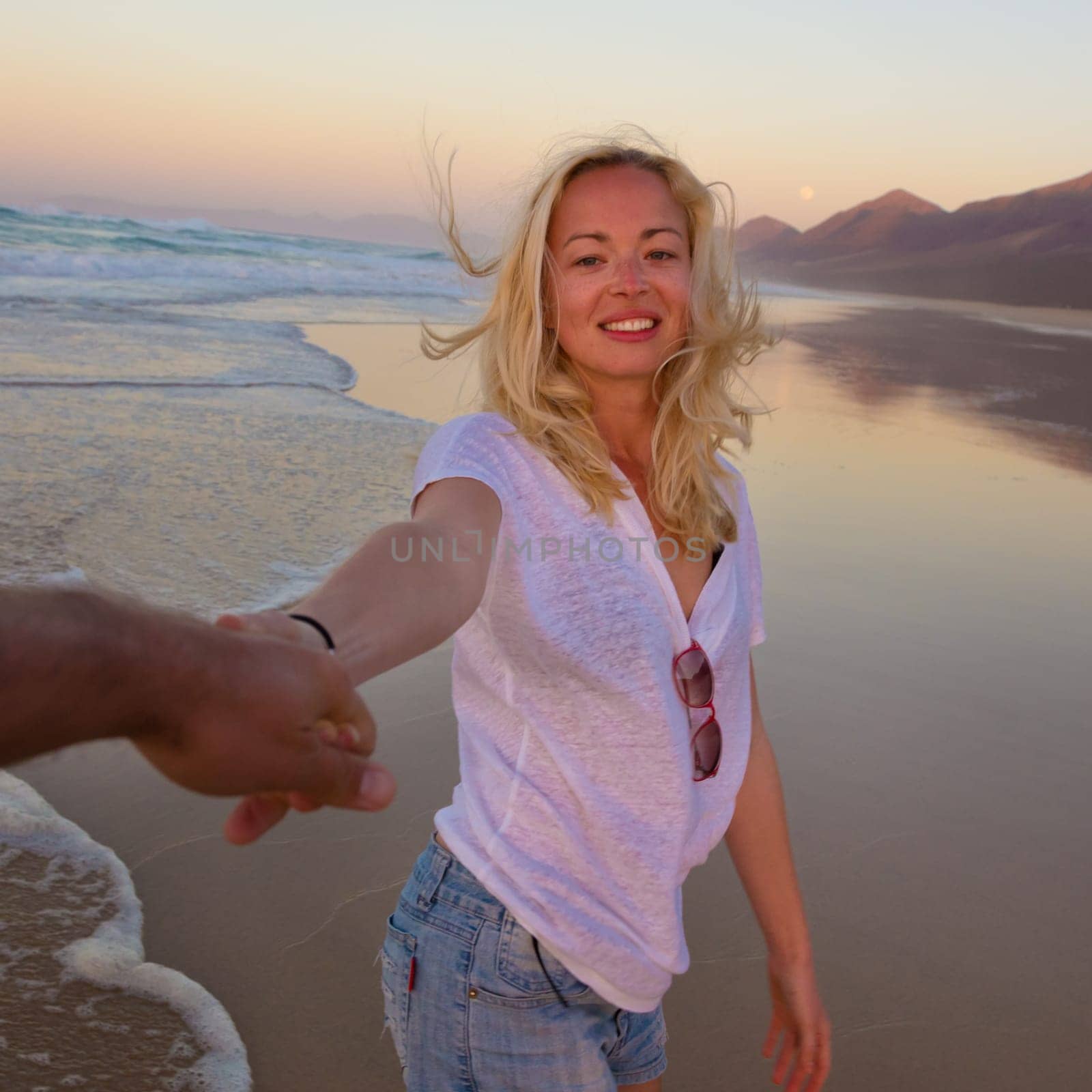 Young romantic couple, holding hands, having fun on perfect deserted beach at sunset. Shot from boyfrieds perspective. Guy looking at her beautiful carefree girlfriend.