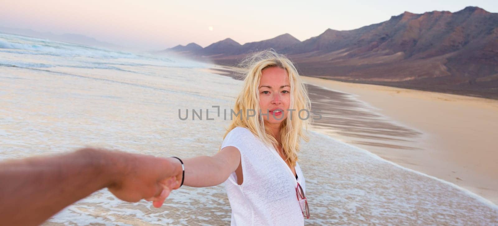Young romantic couple, holding hands, having fun on perfect deserted beach at sunset. Shot from boyfrieds perspective. Guy looking at her beautiful carefree girlfriend.