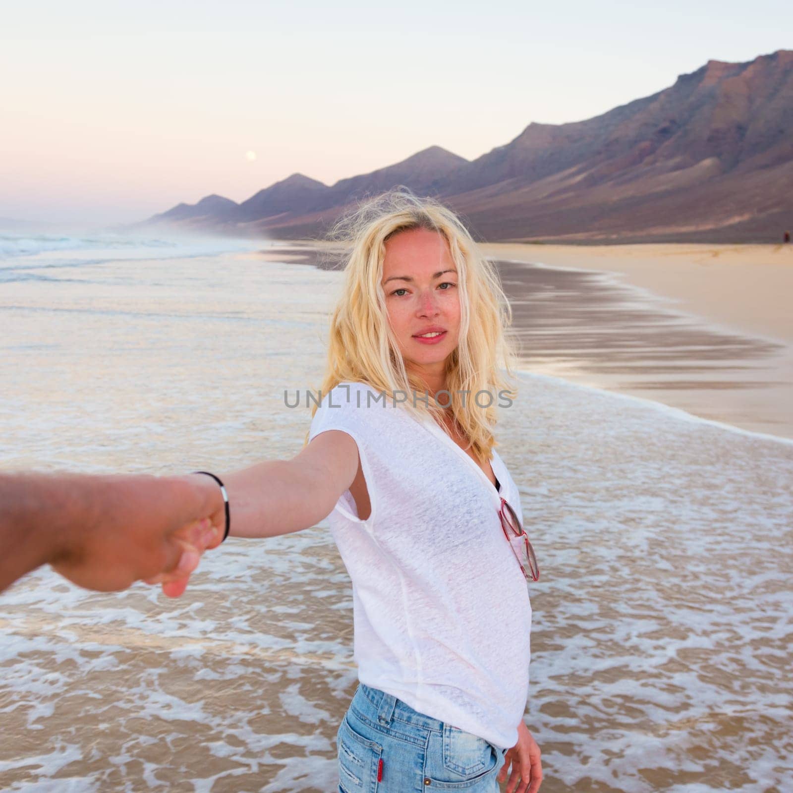 Young romantic couple, holding hands, having fun on perfect deserted beach at sunset. Shot from boyfrieds perspective. Guy looking at her beautiful carefree girlfriend.