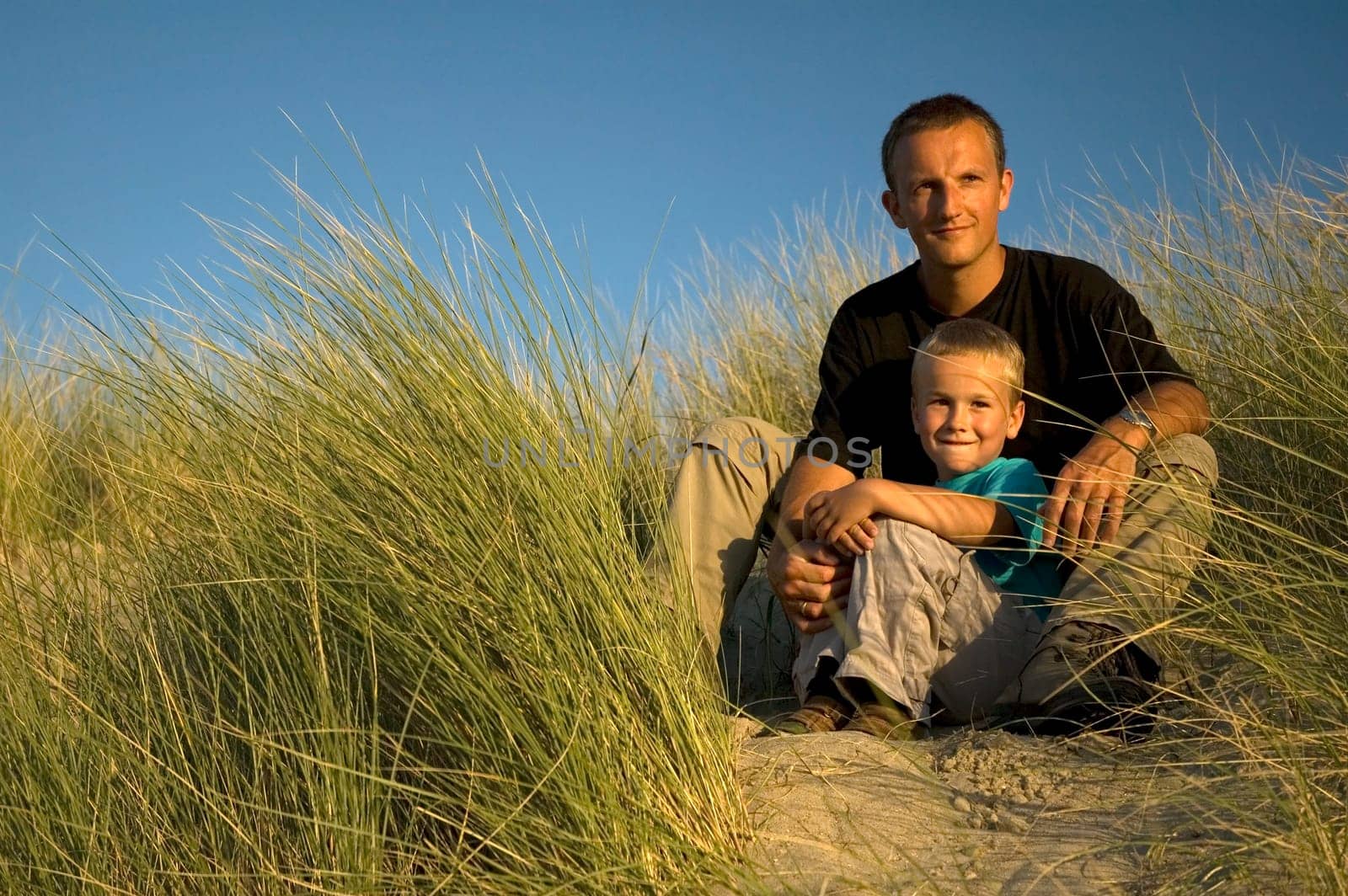 A boy and his father are looking far away, to the horizon. They are sitting in the dunes.