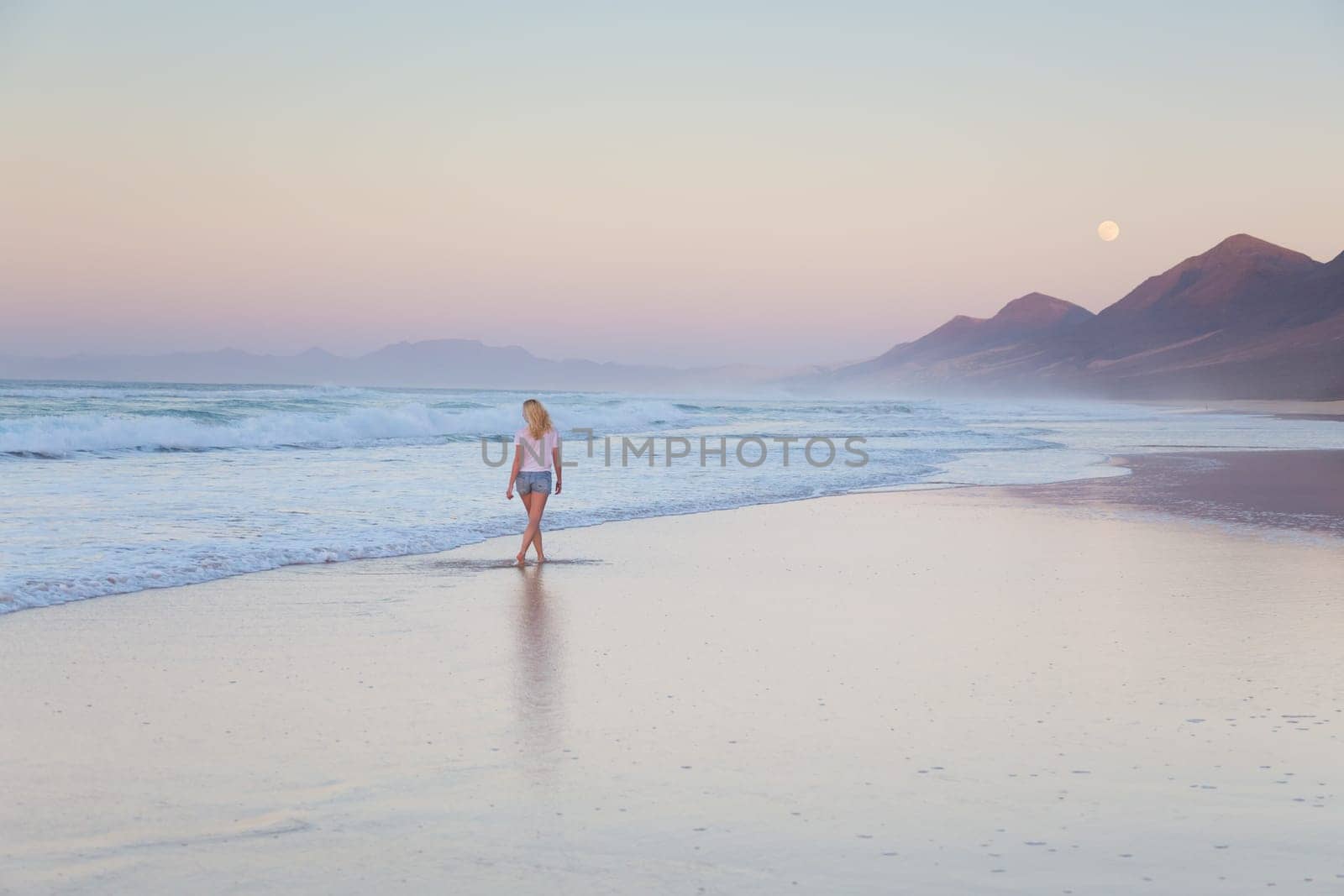 Woman walking on sandy beach in sunset. Waves sweeping away her traces in sand. Beach, travel, concept. Copy space.