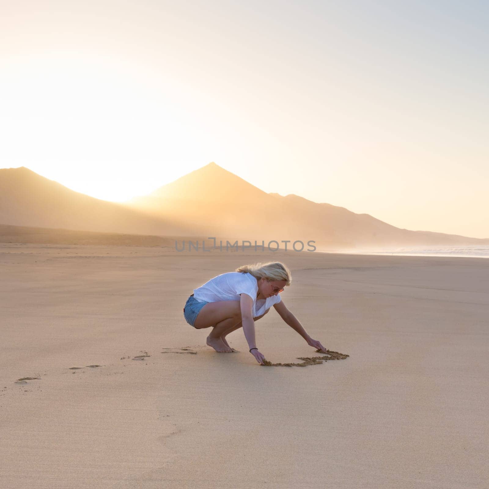 Woman drawing heart in sand on beautiful solitary Cofete beach in sunset. Love, travel concept. Fuerteventura, Canary Islands, Spain.