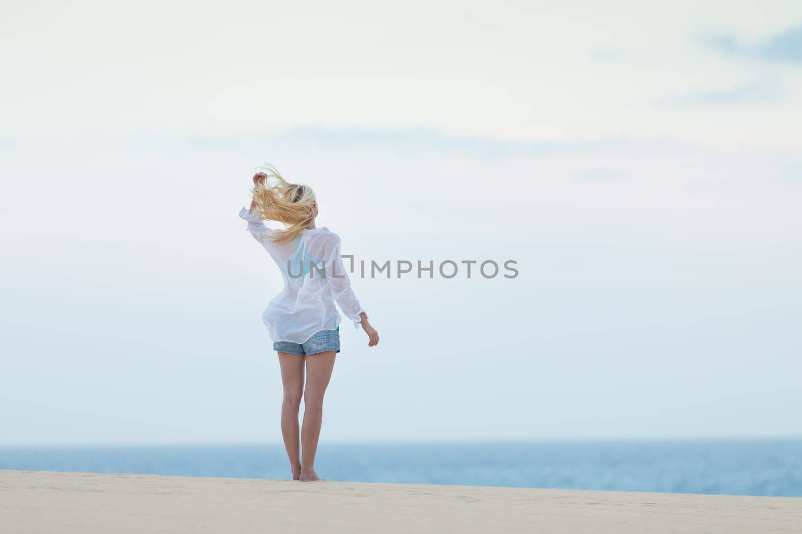 Meditative, sensual blonde woman wearing white loose casual shirt on vacations, looking at horizon, enjoying peaceful early morning.