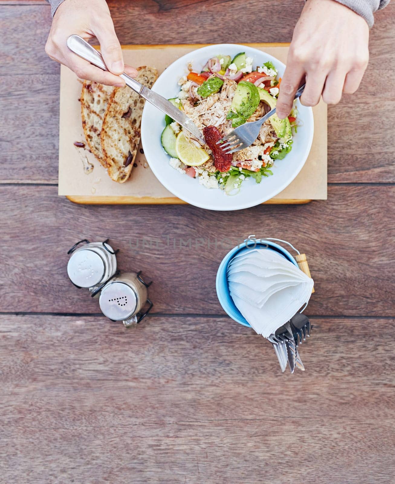 Overhead shot of a person&#39 s hands holding cutlery and about to start eating a fresh salad of chicken, avocado, sundried tomatoes and other ingredients on a wooden table with serviettes at the ready