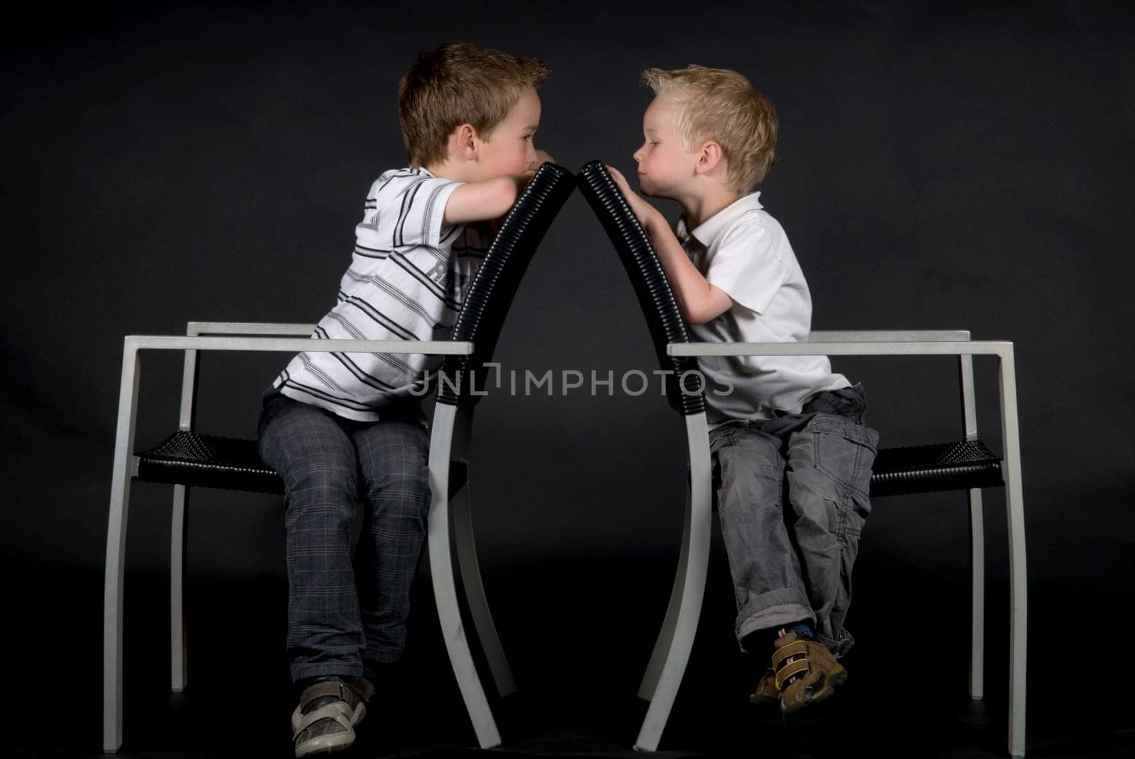 Two brothers sitting on a chair looking at each other, against a black background.