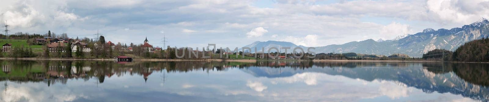 north-eastern panoramic view over the Weissensee near the town of Füssen in the Ostalgäu with beautiful water reflection and the village of Weissensee as well as snow-capped mountains in the background