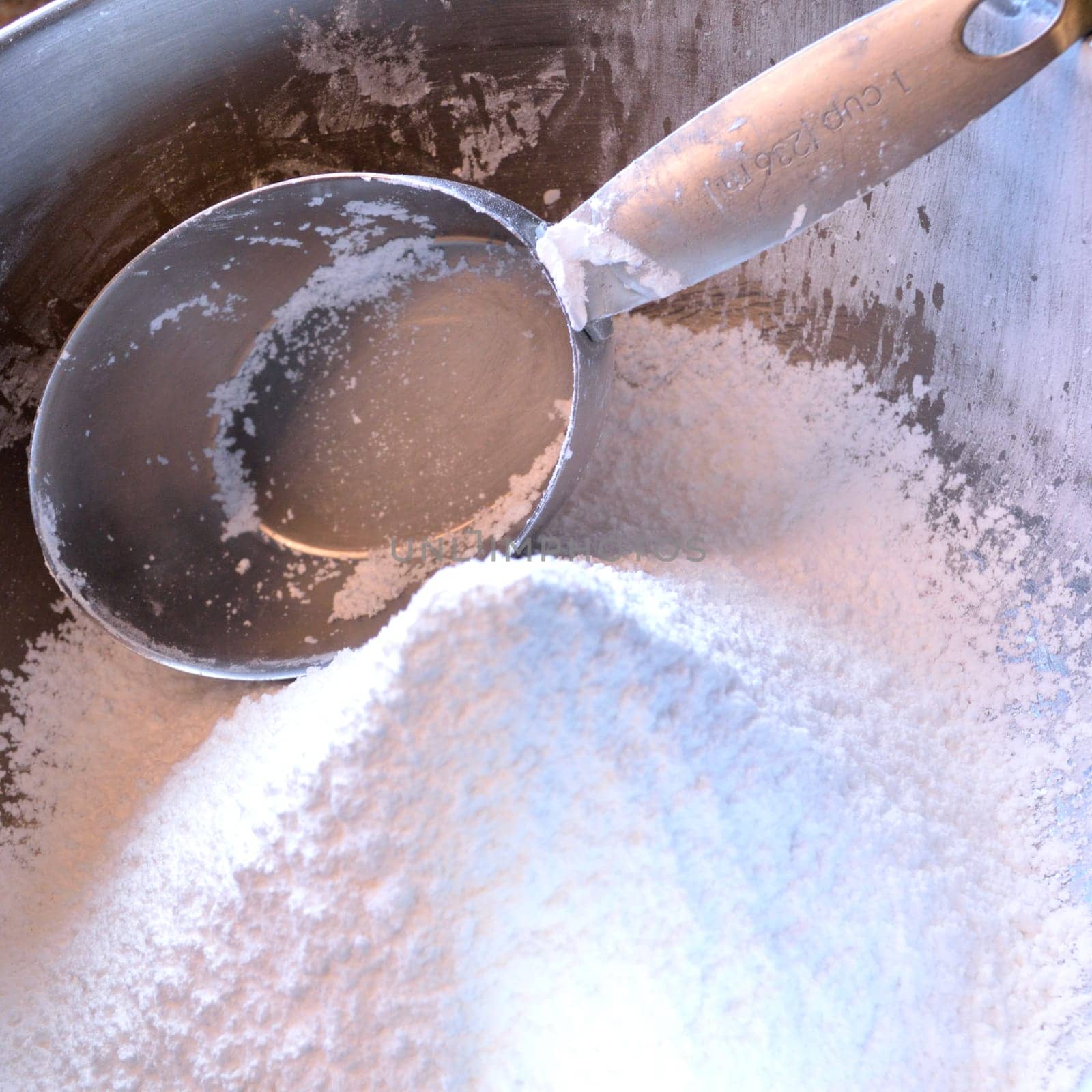 Square photo of a stainless steel measuring cup in a bowl of confectioner's sugar, freshly sifted.