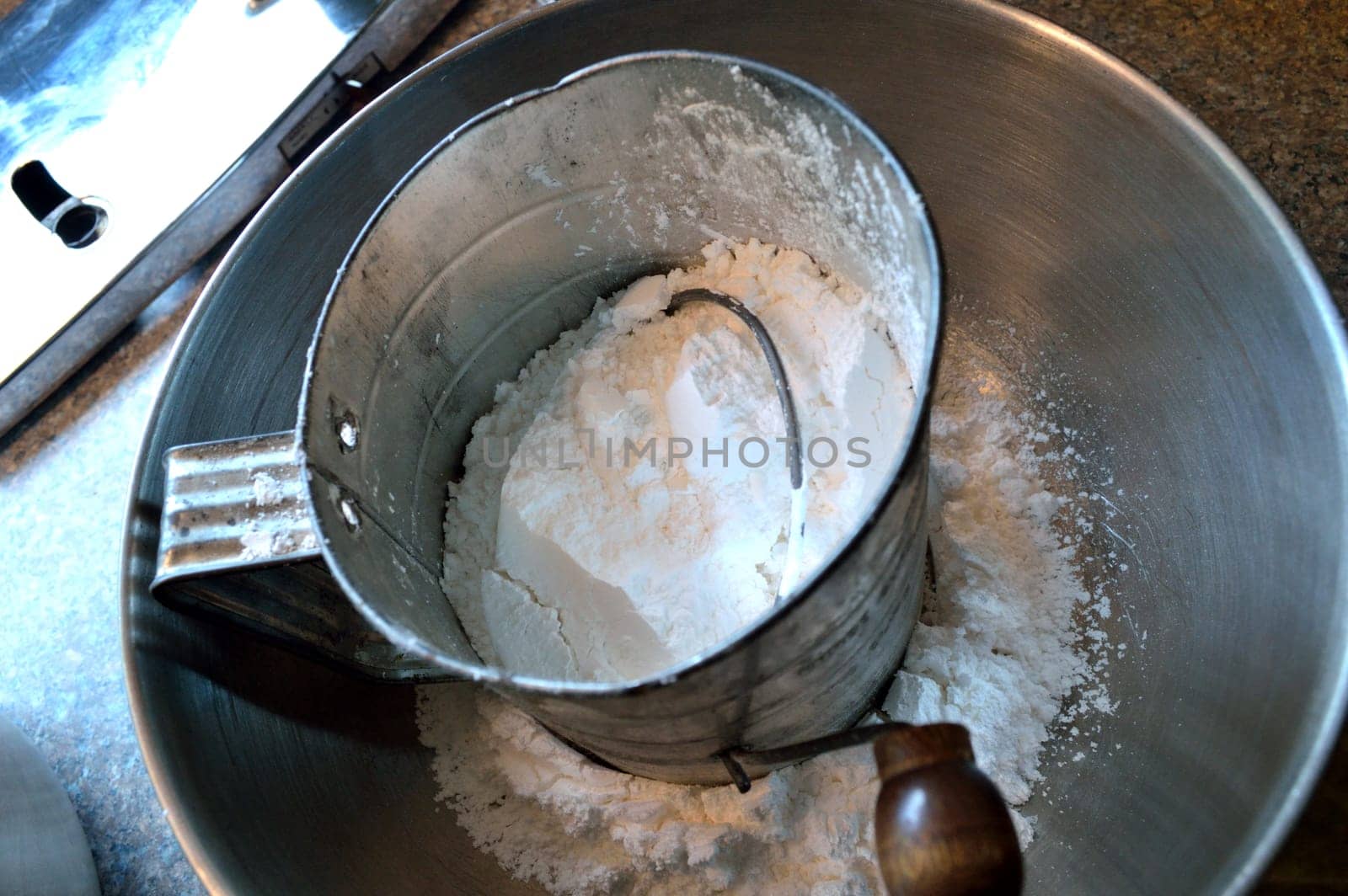 Overhead view of confectioner's (powdered) sugar in an old-fashioned hand sifter in a stainless steel bowl on countertop, with bottom of stand mixer to the left.