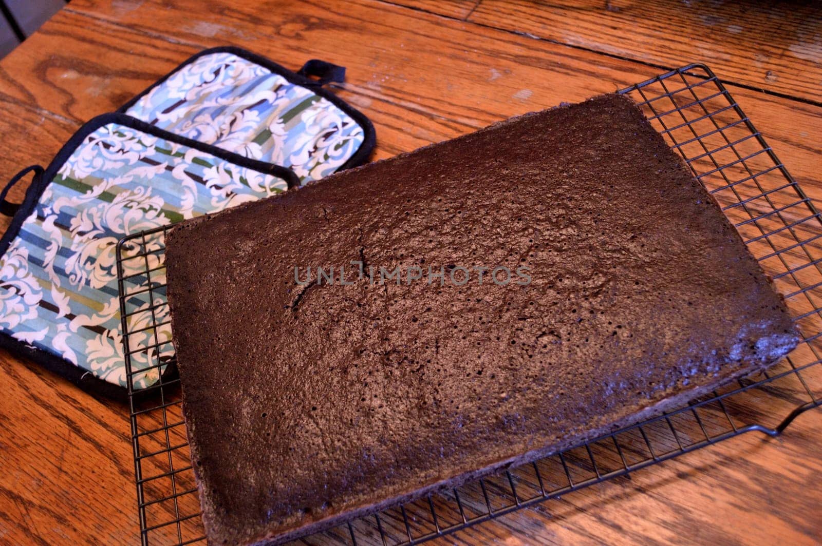 Dark chocolate cake sitting on a cooling rack to cool with colorful oven holders and sea green antique bowl with cake mix in it background on distressed wooden table in old-fashioned kitchen.