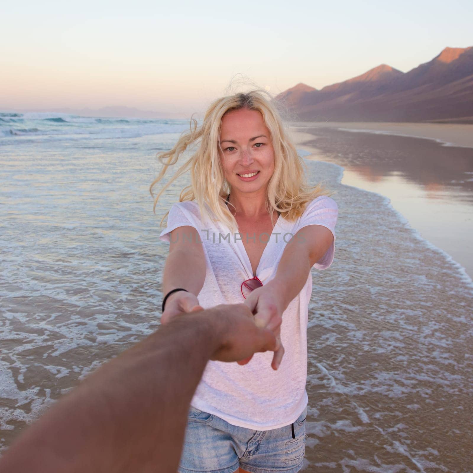Young romantic couple, holding hands, having fun on perfect deserted beach at sunset. Shot from boyfrieds perspective. Guy looking at her beautiful carefree girlfriend.