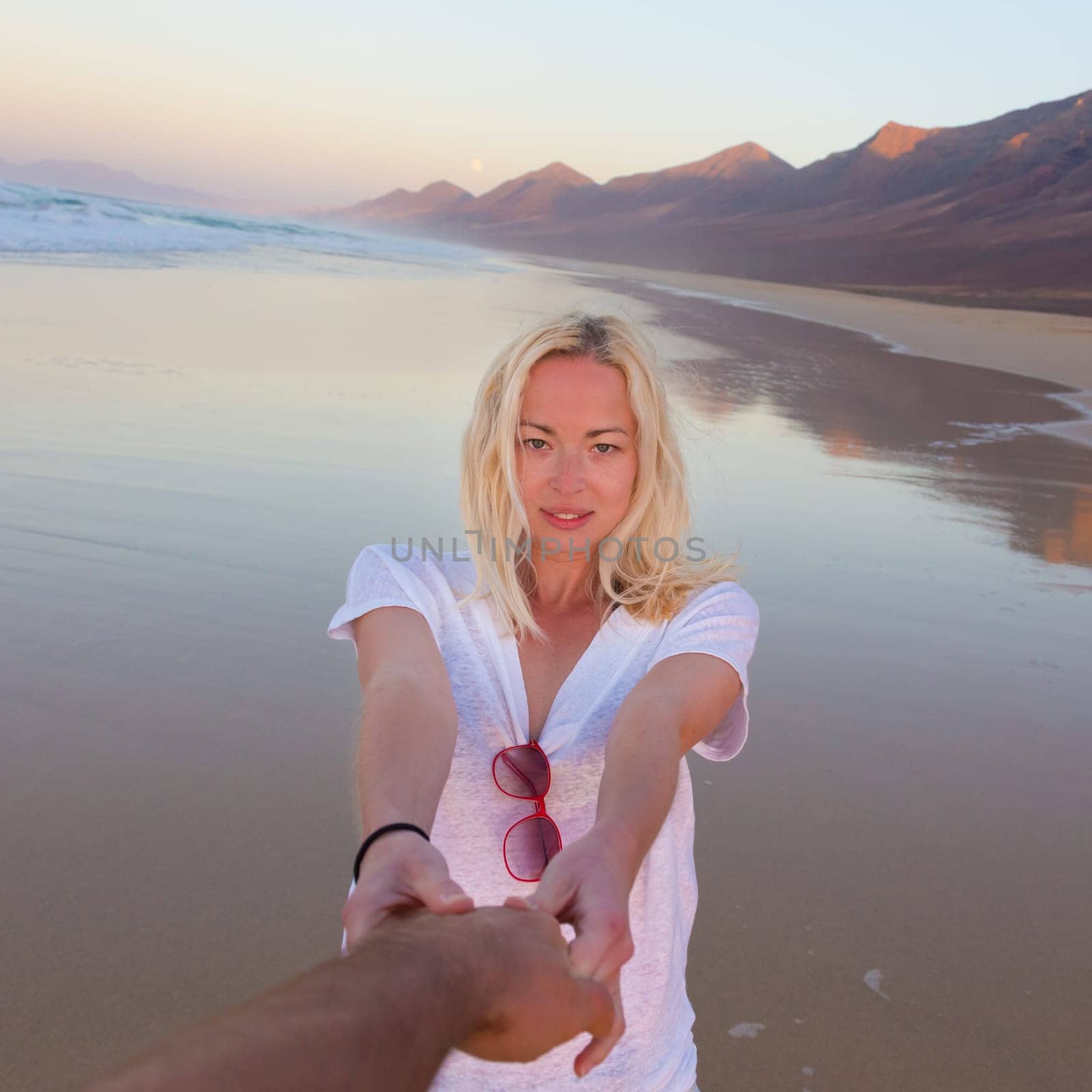 Young romantic couple, holding hands, having fun on perfect deserted beach at sunset. Shot from boyfrieds perspective. Guy looking at her beautiful carefree girlfriend.
