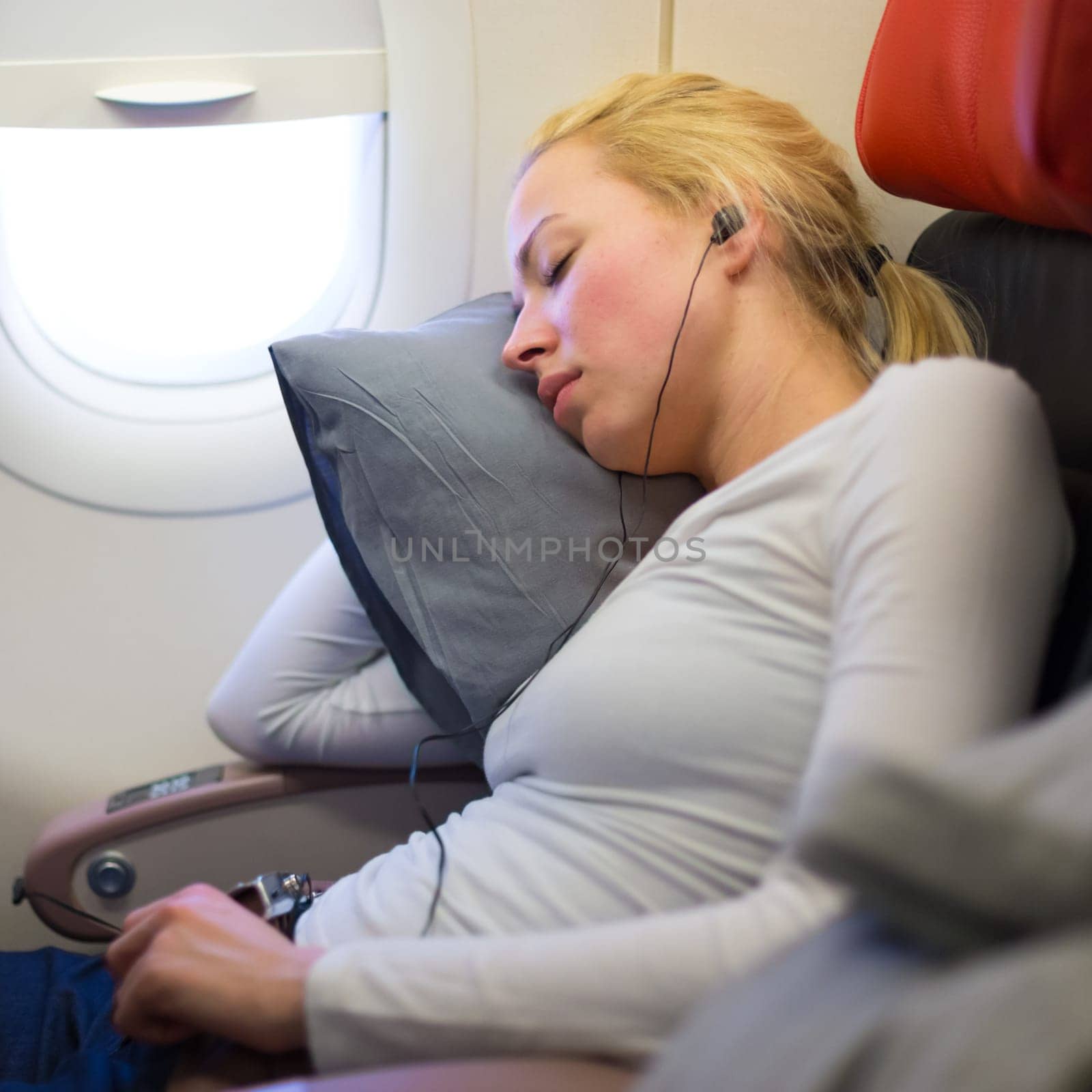 Tired blonde casual caucasian lady listens to music while napping on uncomfortable seat while traveling by airplane. Commercial transportation by planes.