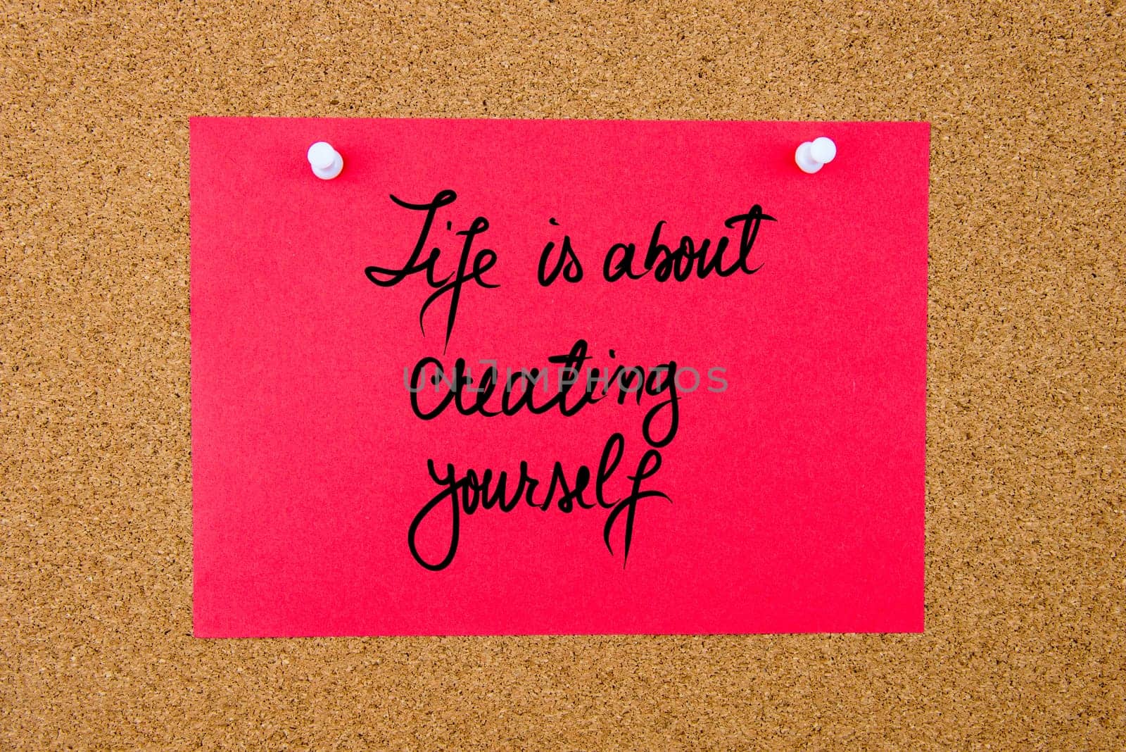 Red paper note with handwritten text Life Is About Creating Yourself pinned on cork board with white thumbtacks