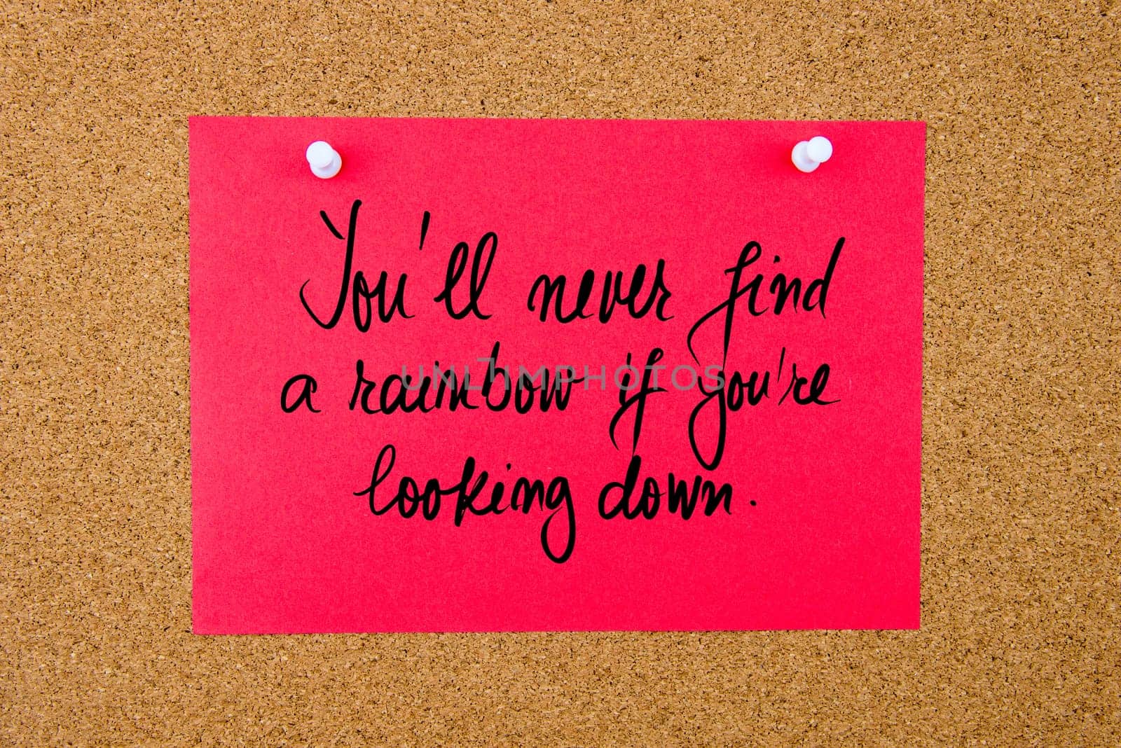 Red paper note with handwritten text You Will Never Find A Rainbow If You Are Looking Down pinned on cork board with white thumbtacks