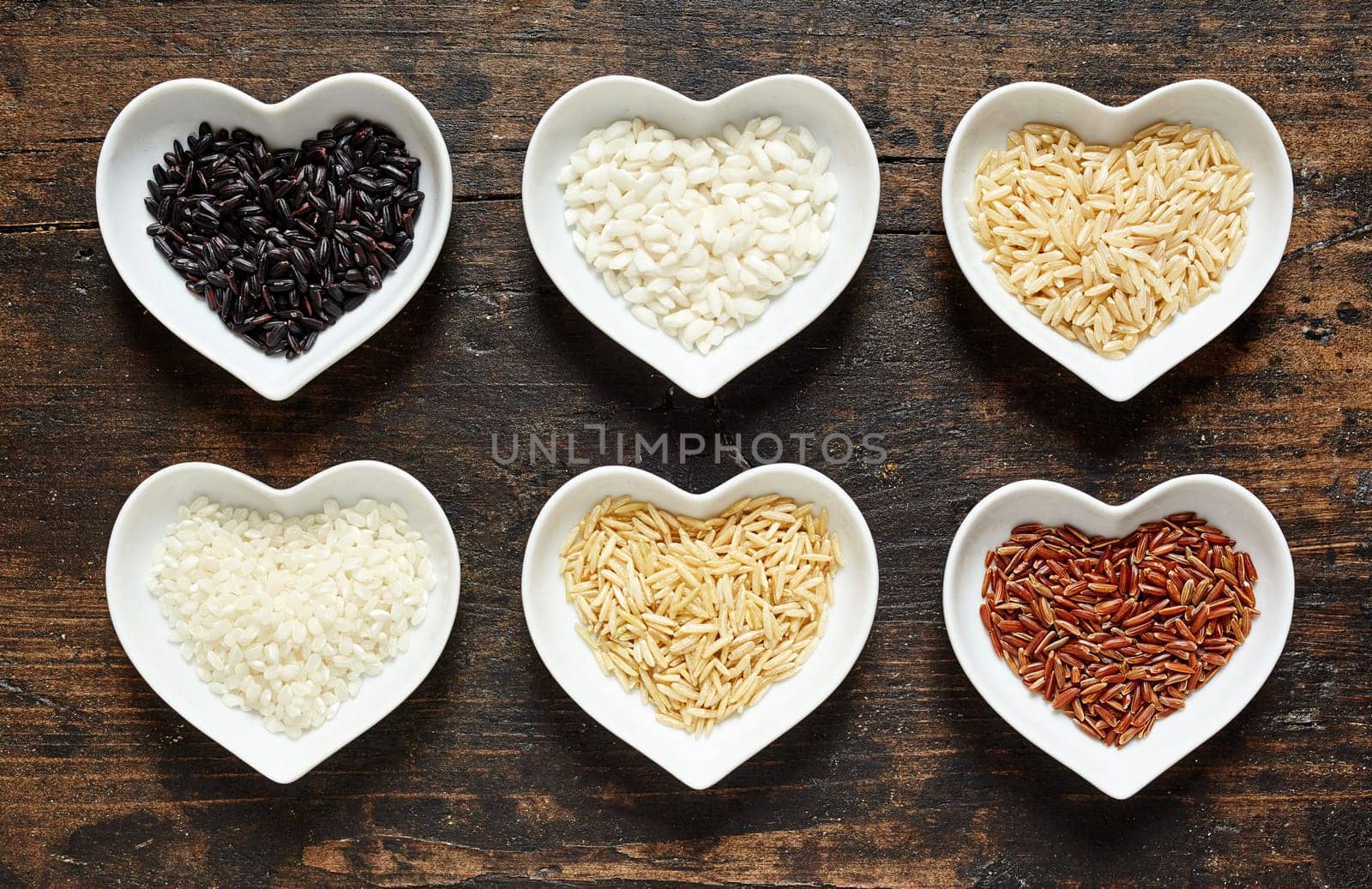 Selection of different rice grains in heart shaped dishes with par-boiled and husked white rice and long grain wild rice, overhead view on rustic wood