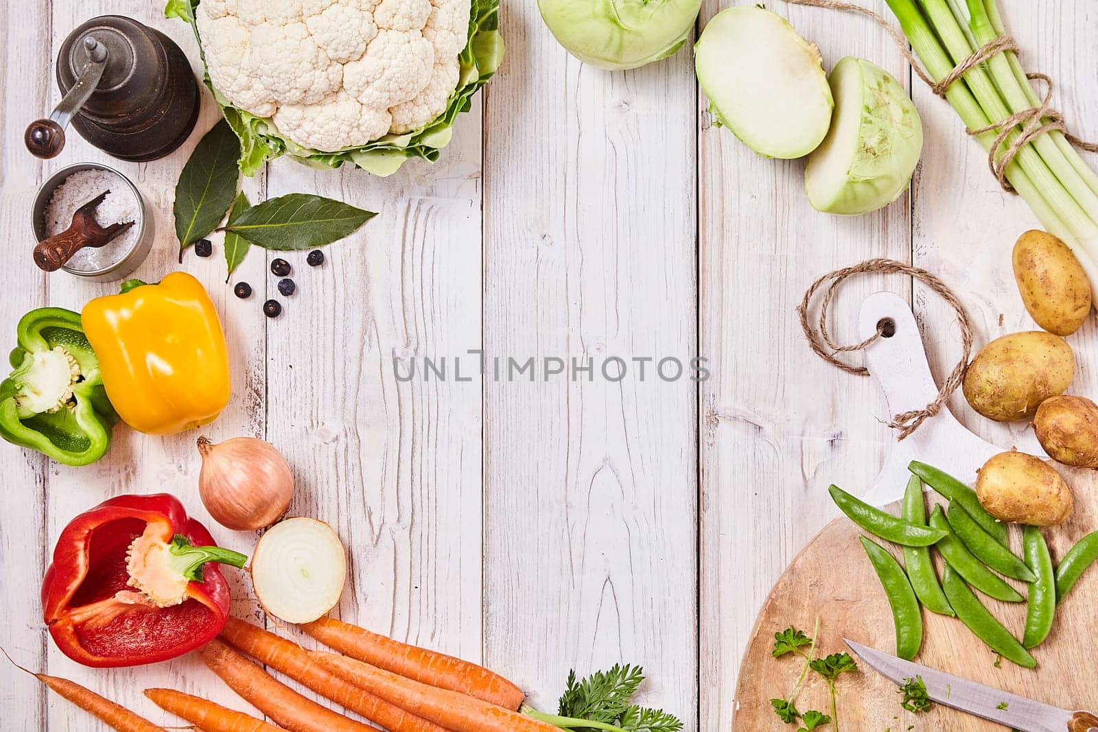 Delicious fresh vegetables forming a frame on white wood with central copy space together with a cutting board, knife and seasoning for cooking