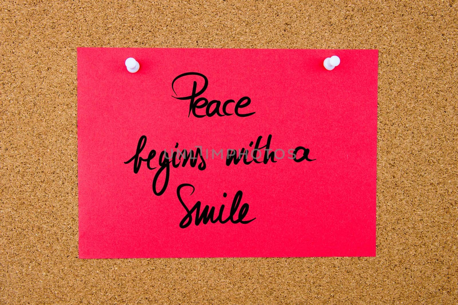 Red paper note with handwritten text Peace Begins With A Smile pinned on cork board with white thumbtacks