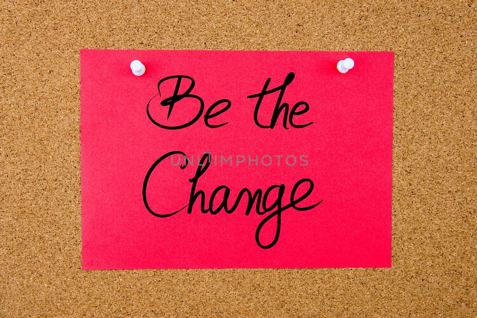 Red paper note with handwritten text Be The Change pinned on cork board with white thumbtacks