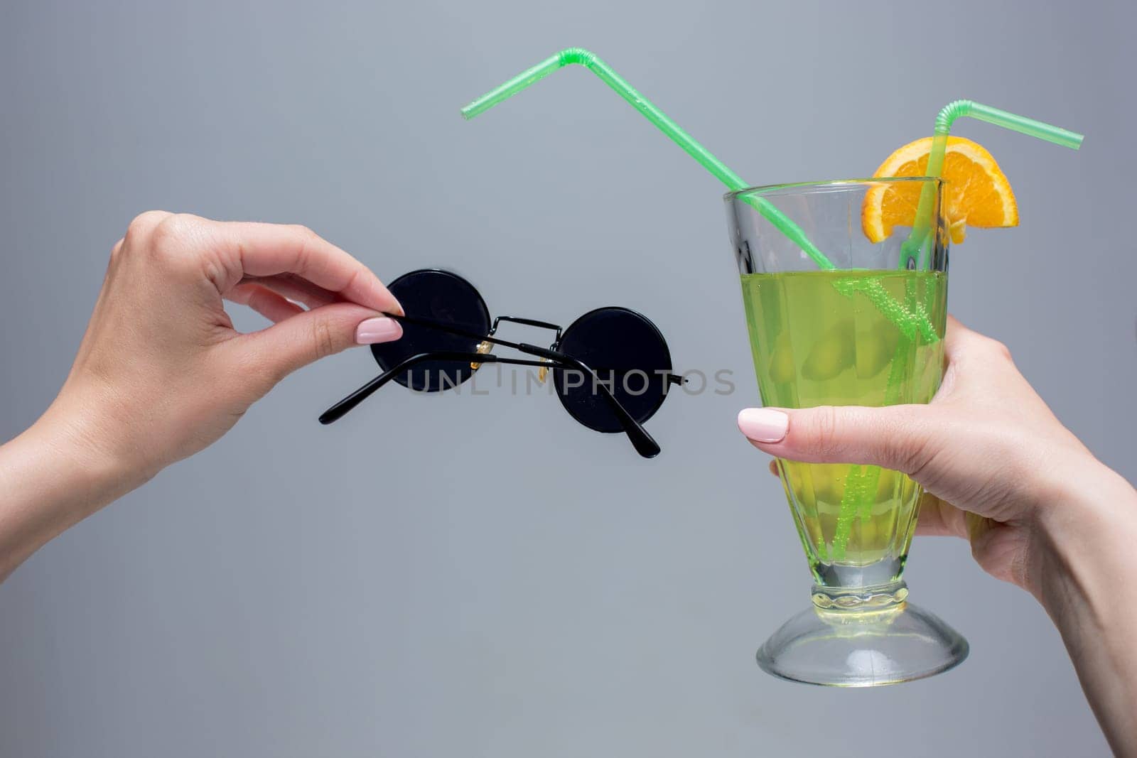 The female hands with cocktail and sunglasses on a gray background