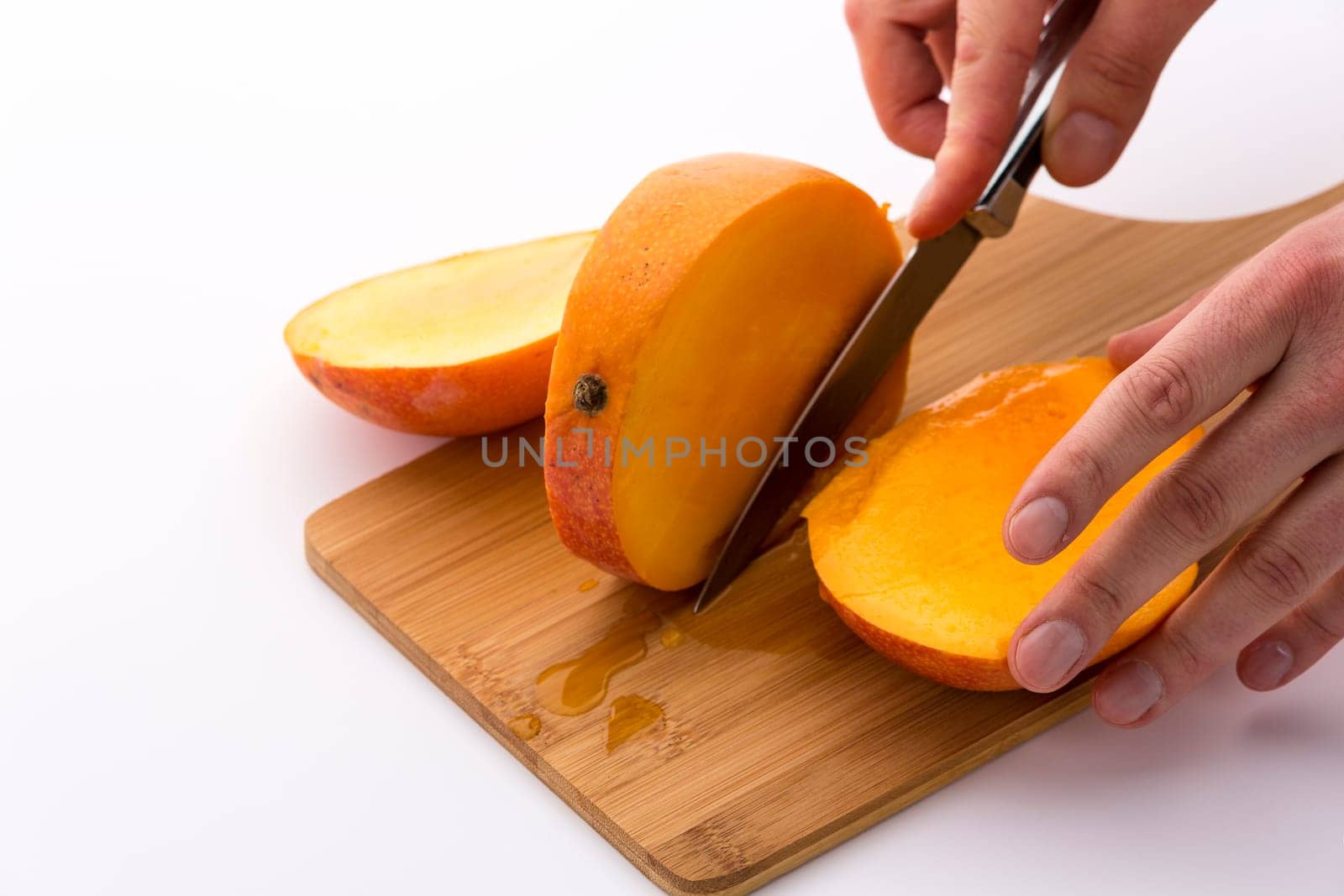 Trisecting a mango with a second cut along its oblong, flat pit. Finger tips exerting gentle pressure onto the kitchen knife used. Yellow and tangerine fruit pulp in orange skin. White background.