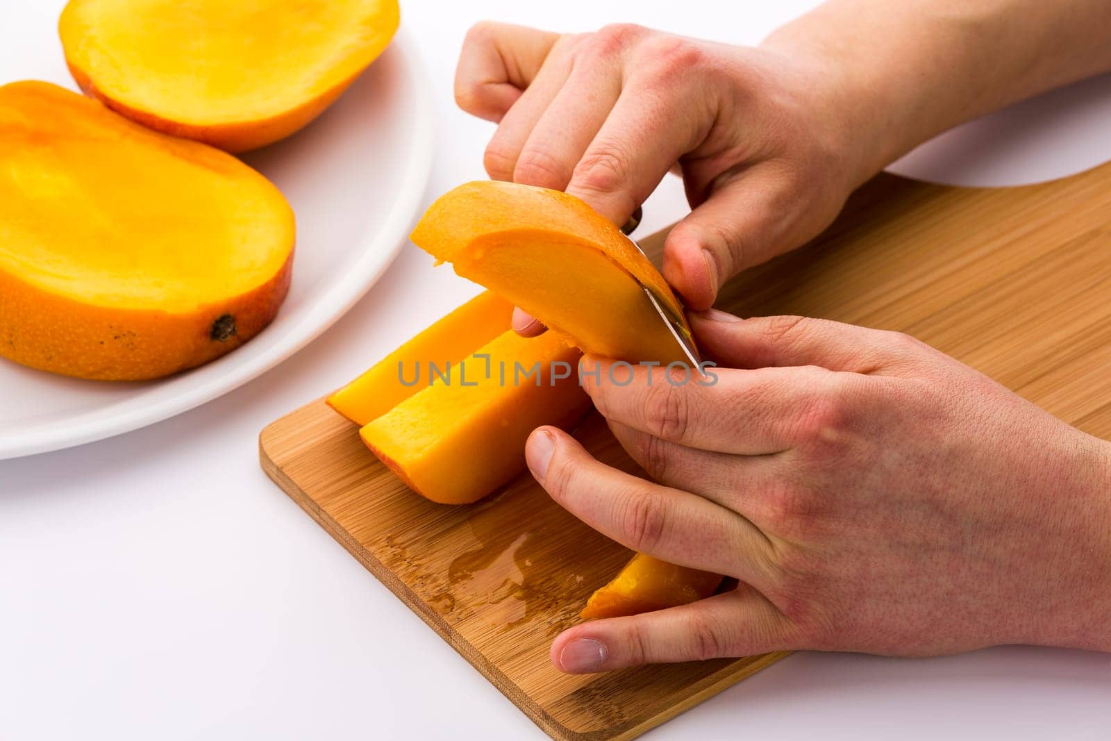 Closeup of a man’s hands peeling one of four mango wedges over a bamboo cutting board. The blade of a short kitchen knife is separating the yellow, juicy fruit pulp from its skin. White background.
