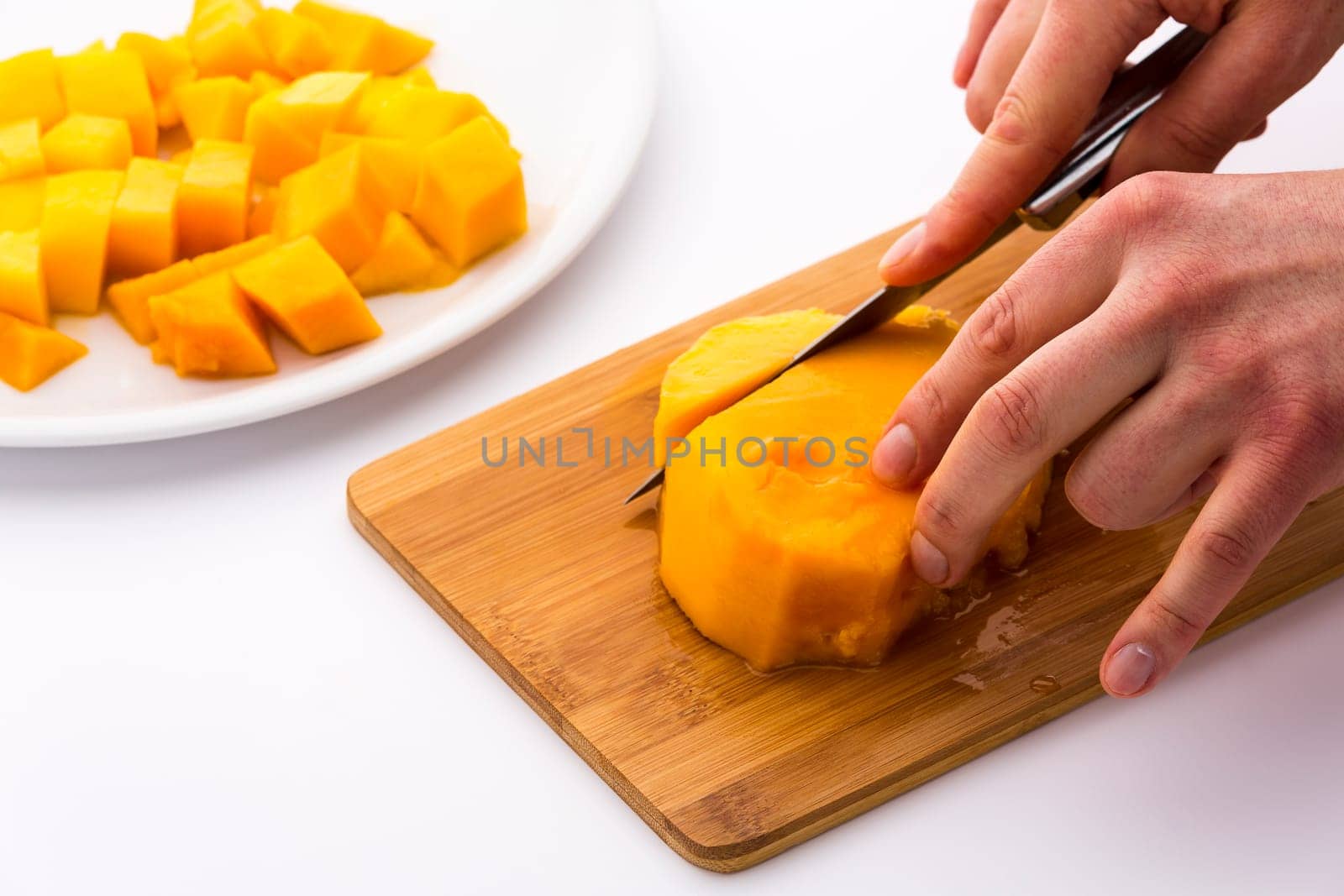 First cut through the middle third of a mango containing its fruit pit. Left finger tips are placing the juicy mango flesh on a wooden board, while a right hand is separating a pulp chip with a knife.
