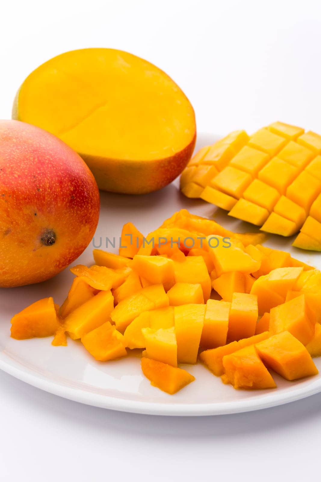 Heap of freshly cut mango pulp cubes on a plate. A mango third scored to shape dice that still are attached to the peel. A mango cross-sectioned along its pit. White background. Vertical orientation.