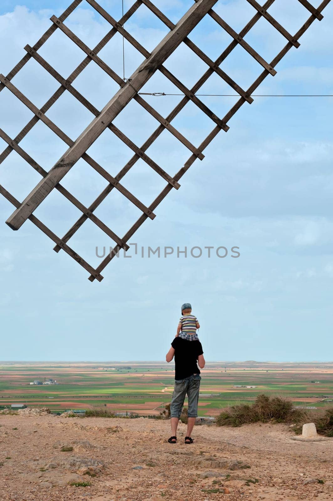 Wooden wing from old windmill over blue sky. Father and his son sitting on father&#39 s shoulders standing turned away