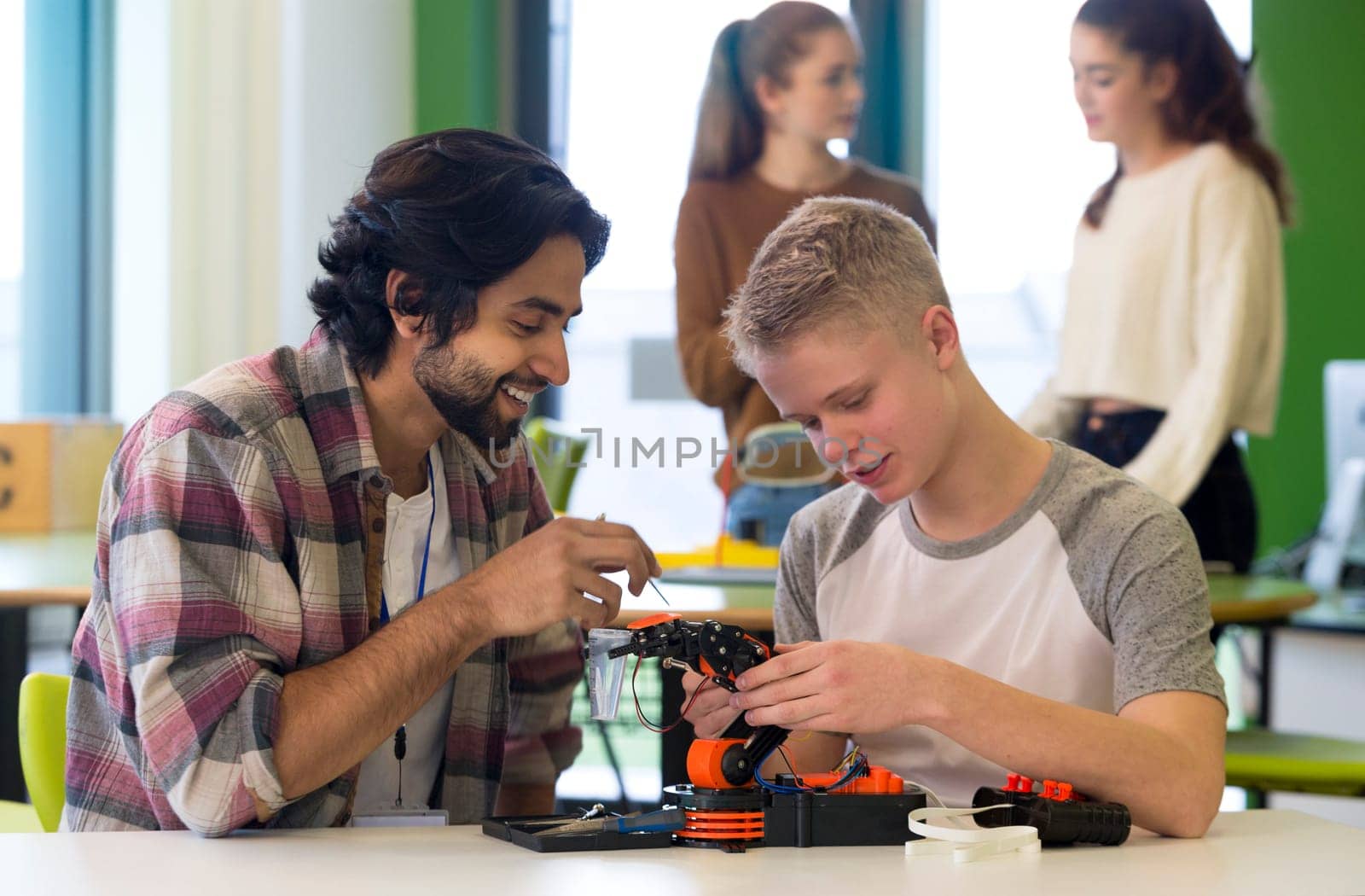Student and teacher working on a robotic arm in a classroom. There are students standing in the background.