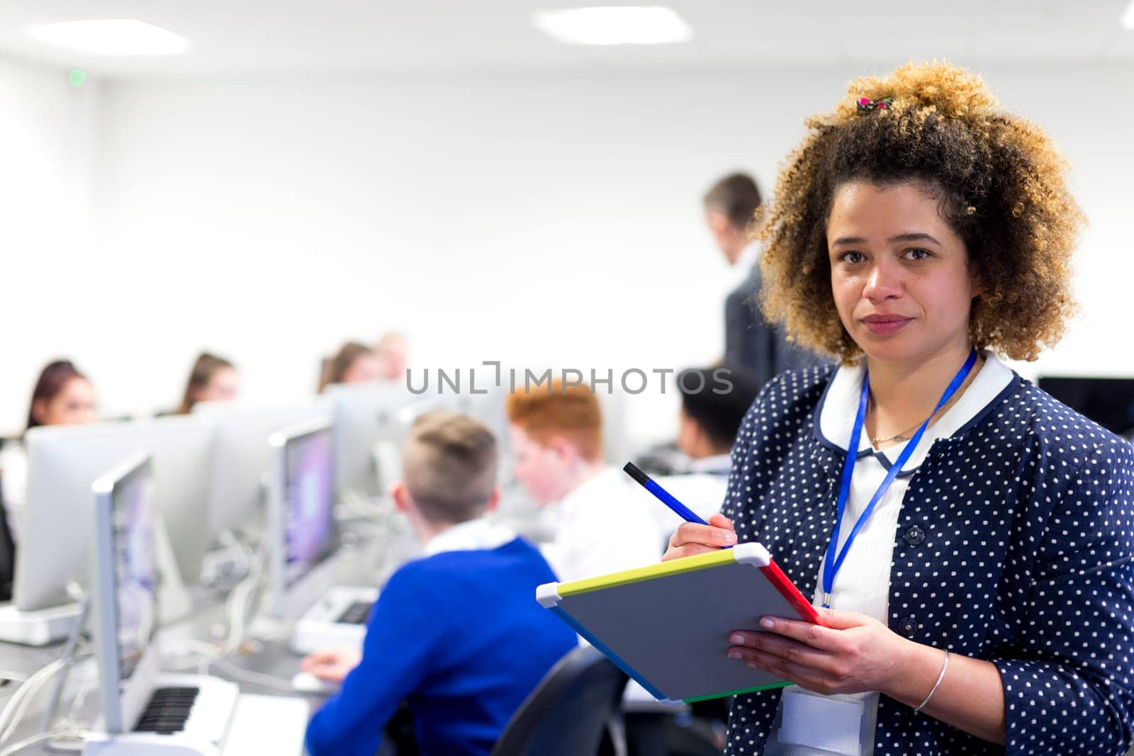 Teacher standing in a classroom with a whiteboard and pen. She is looking at the camera. Students are working on computers in the background.