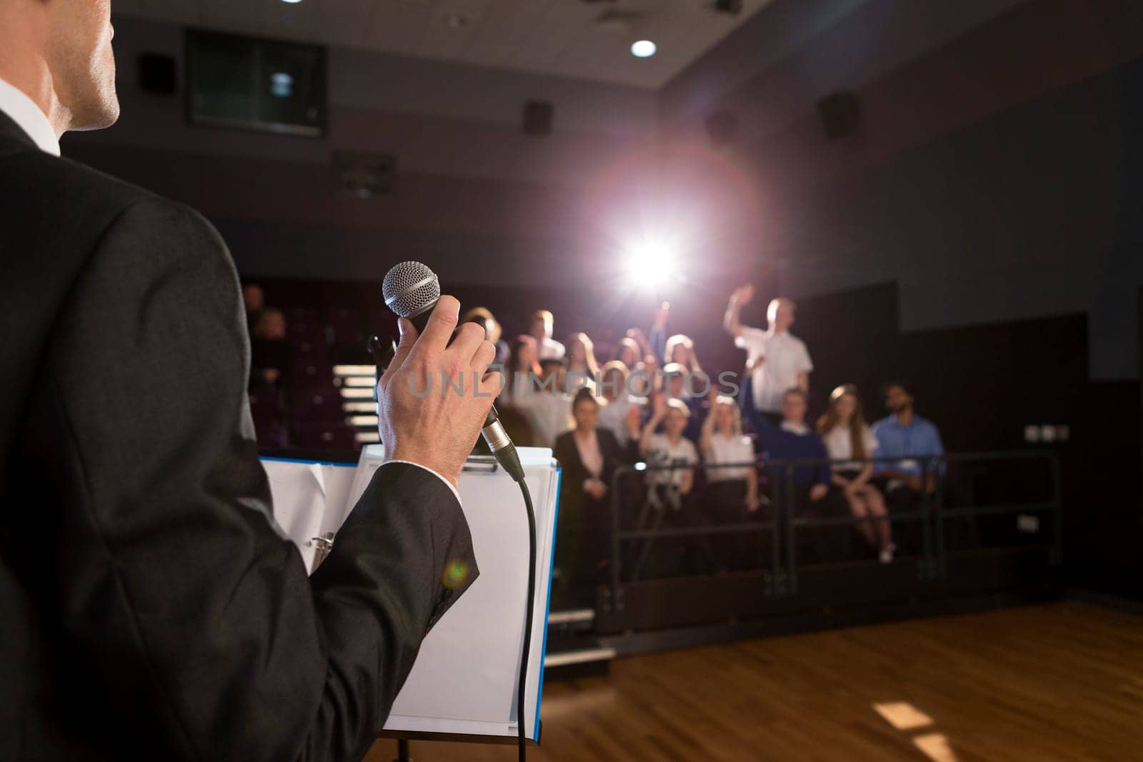Male teacher making a speech in a lecture hall. He is standing at the podium and some students have their hand up to ask a question.