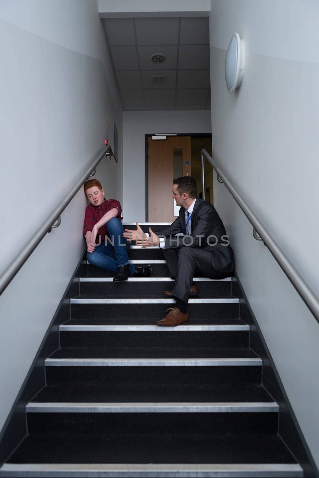 Male student is sitting on the stairs in his school, with his teacher next to him. He is telling him off for his behaviour.
