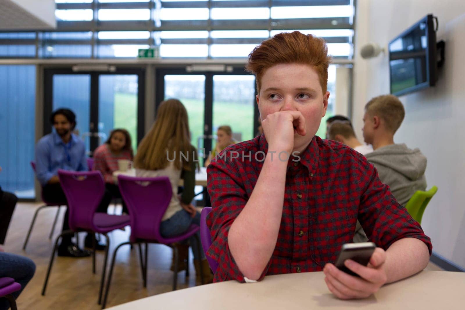 Stressed schoolboy sitting away from people at school. He has a smartphone in his hand and is looking away with his hand to his face.