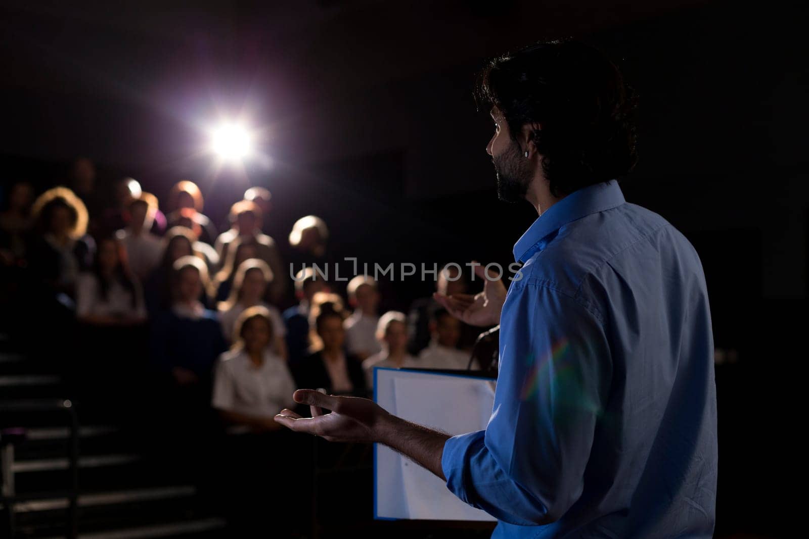 Male teacher giving a speech in a lecture hall to students and teachers.