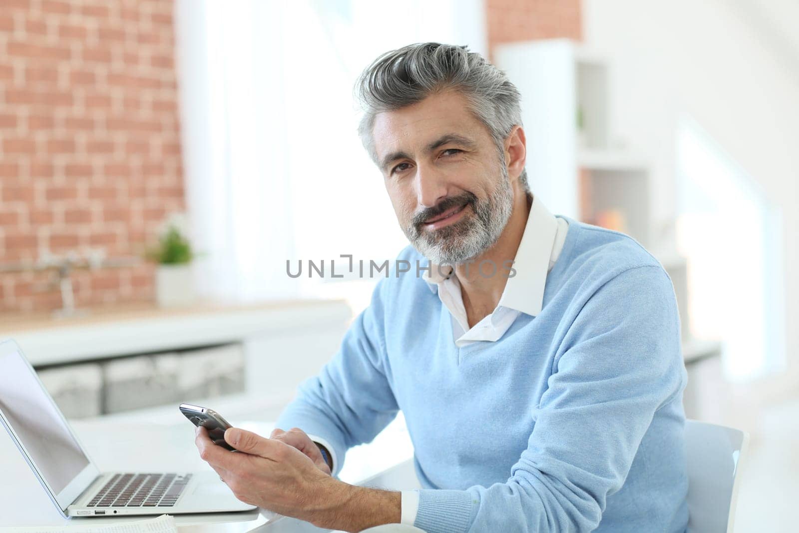 Trendy mature man using smartphone in front of laptop