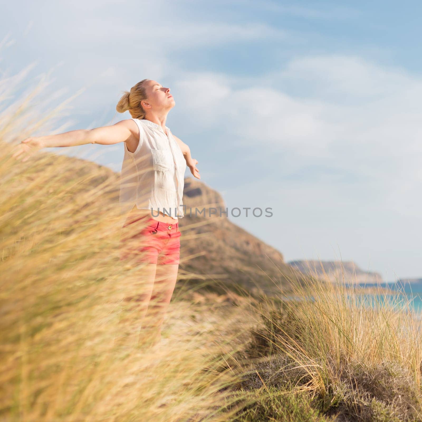 Relaxed woman, arms rised, enjoying sun, freedom and life an a beautiful beach. Young lady feeling free, relaxed and happy. Concept of vacations, freedom, happiness, enjoyment and well being.