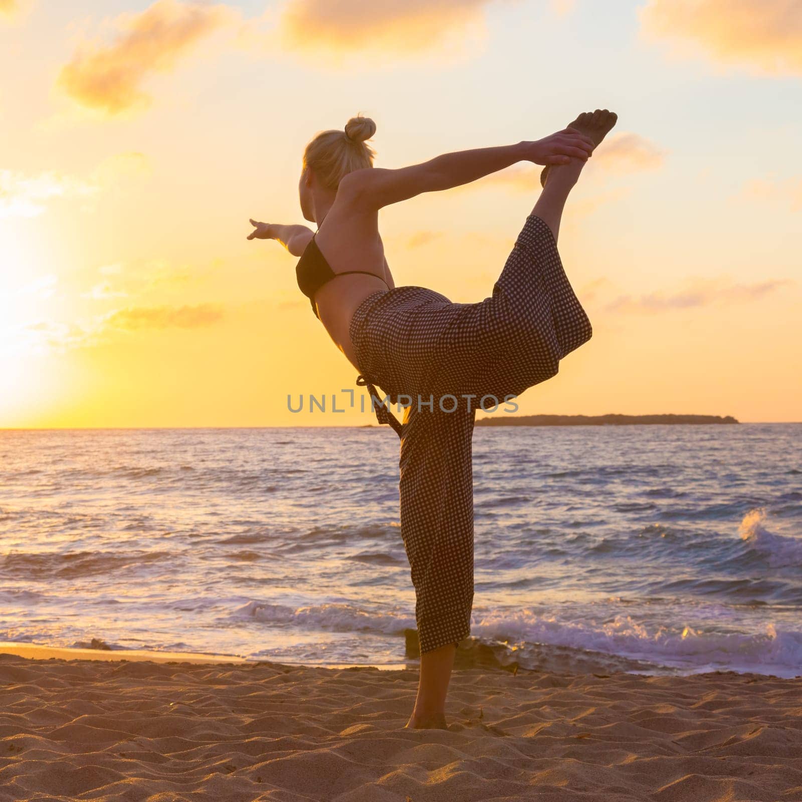 Silhouette of young woman practicing standing bow yoga pose on sandy beach at sunset.