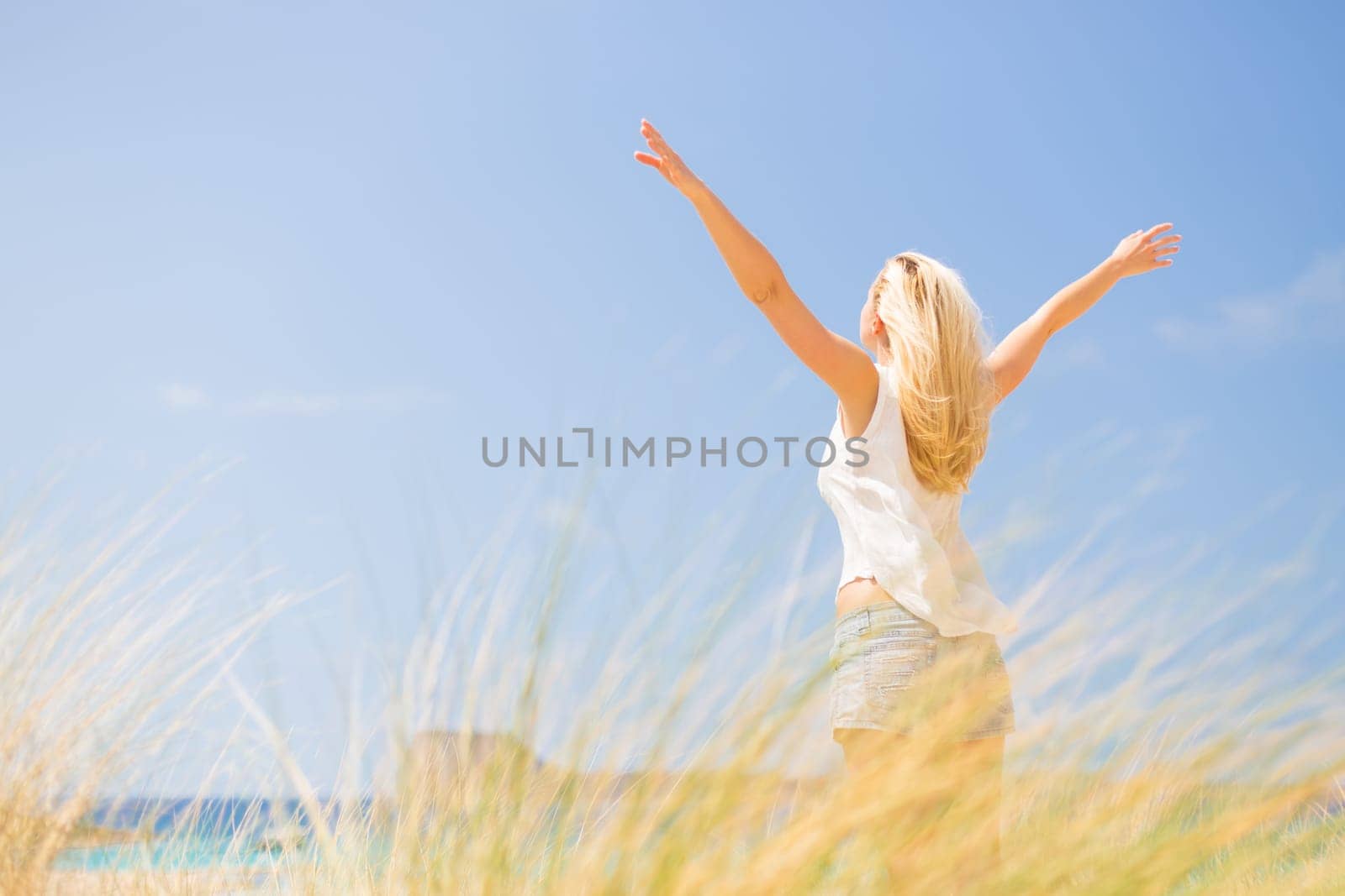 Relaxed woman, arms rised, enjoying sun, freedom and life an a beautiful beach. Young lady feeling free, relaxed and happy. Concept of vacations, freedom, happiness, enjoyment and well being.