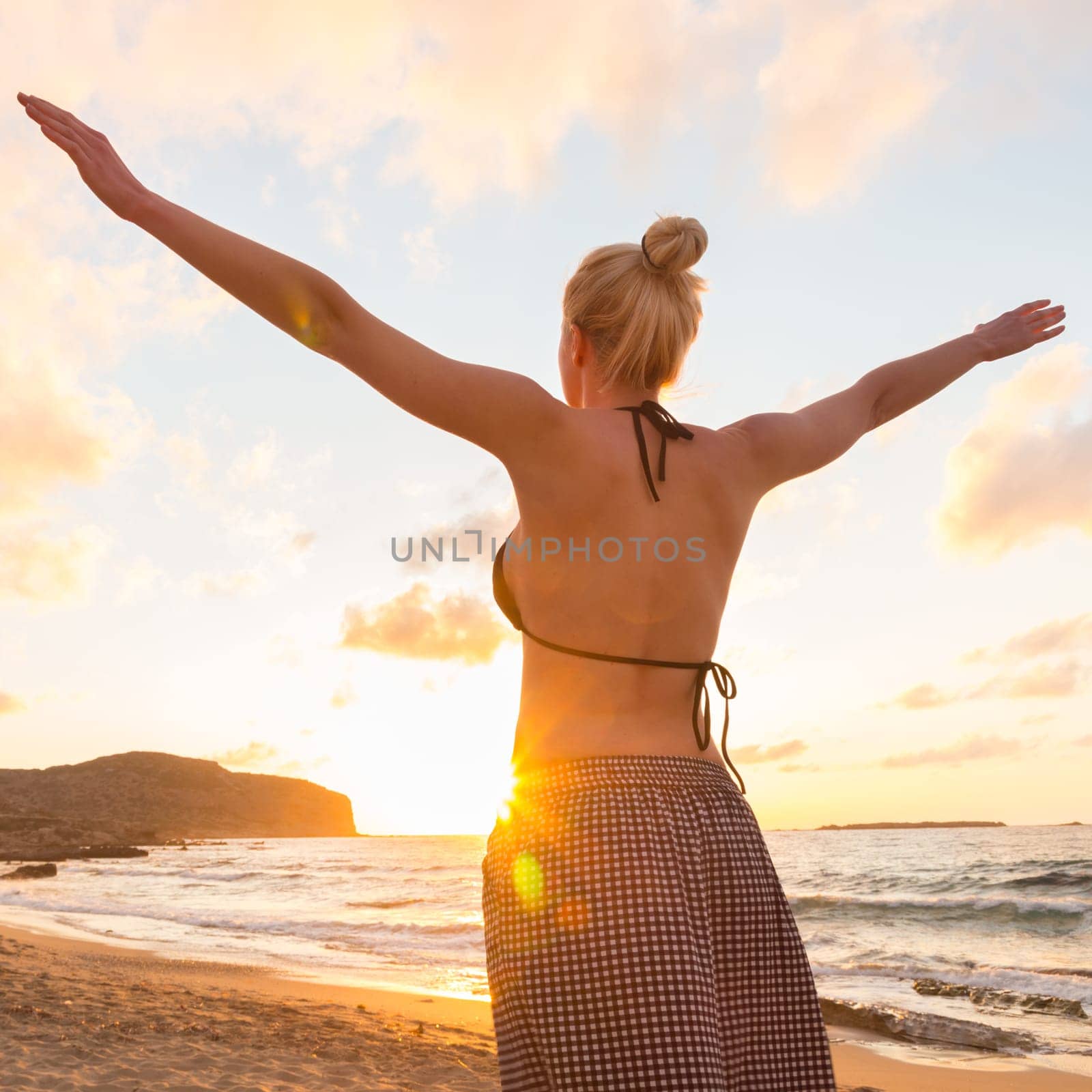 Relaxed woman, arms rised, enjoying sun, freedom and life an beautiful beach in sunset. Young lady feeling free, relaxed and happy. Concept of vacations, freedom, happiness, enjoyment and well being.