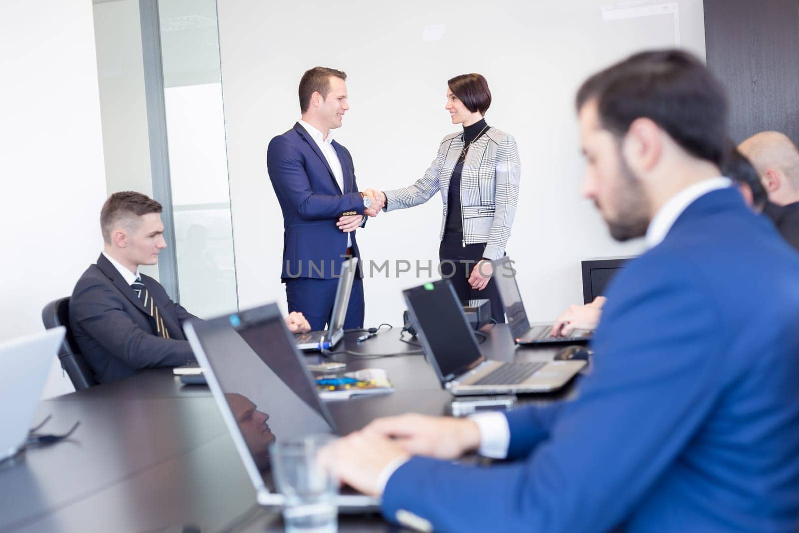 Sealing a deal. Business people shaking hands, finishing up meeting in corporate office. Businessman working on laptop in foreground. Business and entrepreneurship concept.