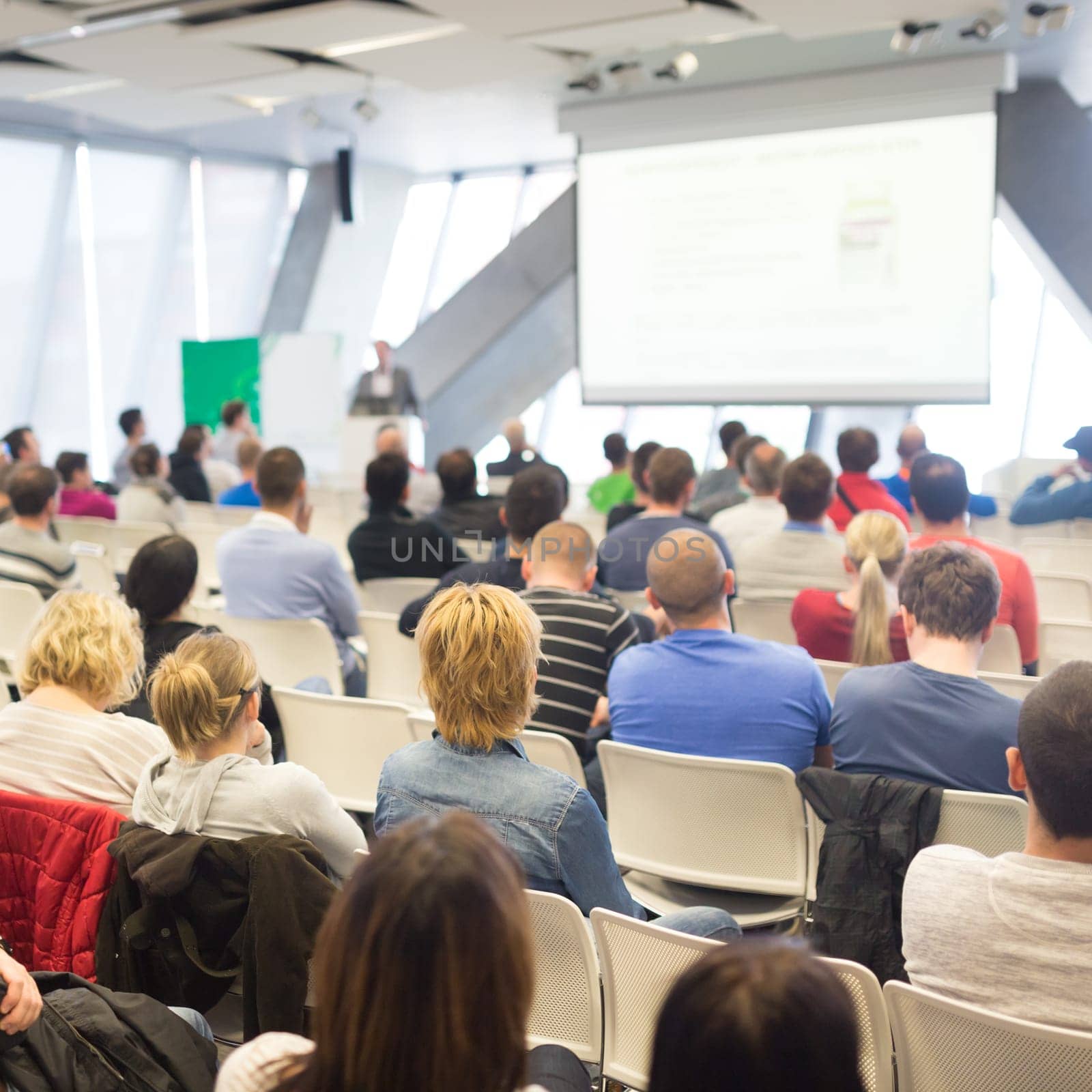 Man giving presentation in lecture hall. Male speeker having talk at public event. Participants listening to lecture. Rear view, focus on people in audience.