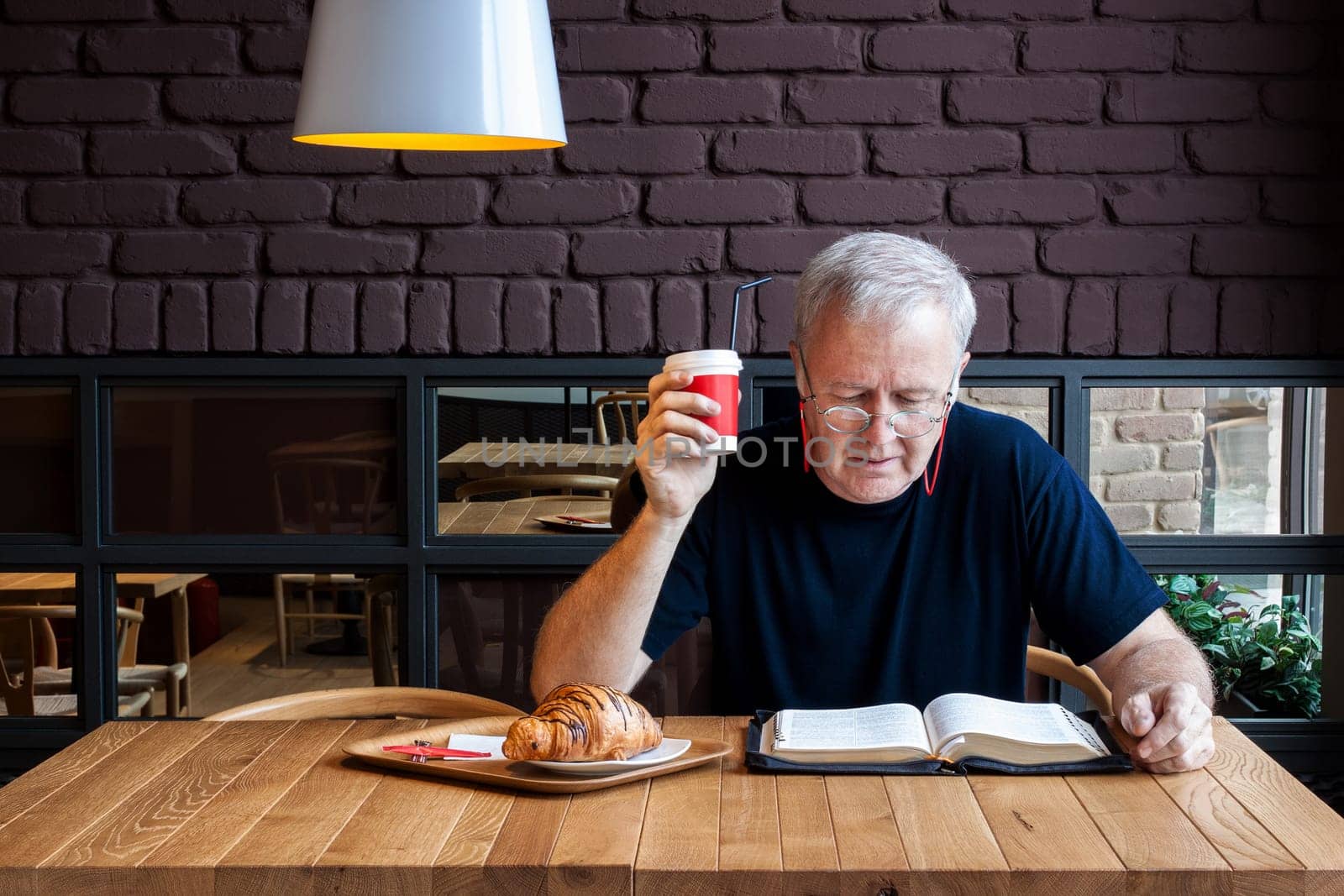 Man sitting at a table with a coffee and a croissant in a snack bar and reading the Bible. Working break dedicated to material and spiritual feeding.