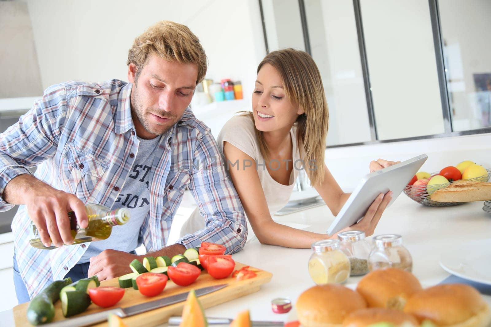 Cheerful couple in kitchen cooking dinner, using tablet