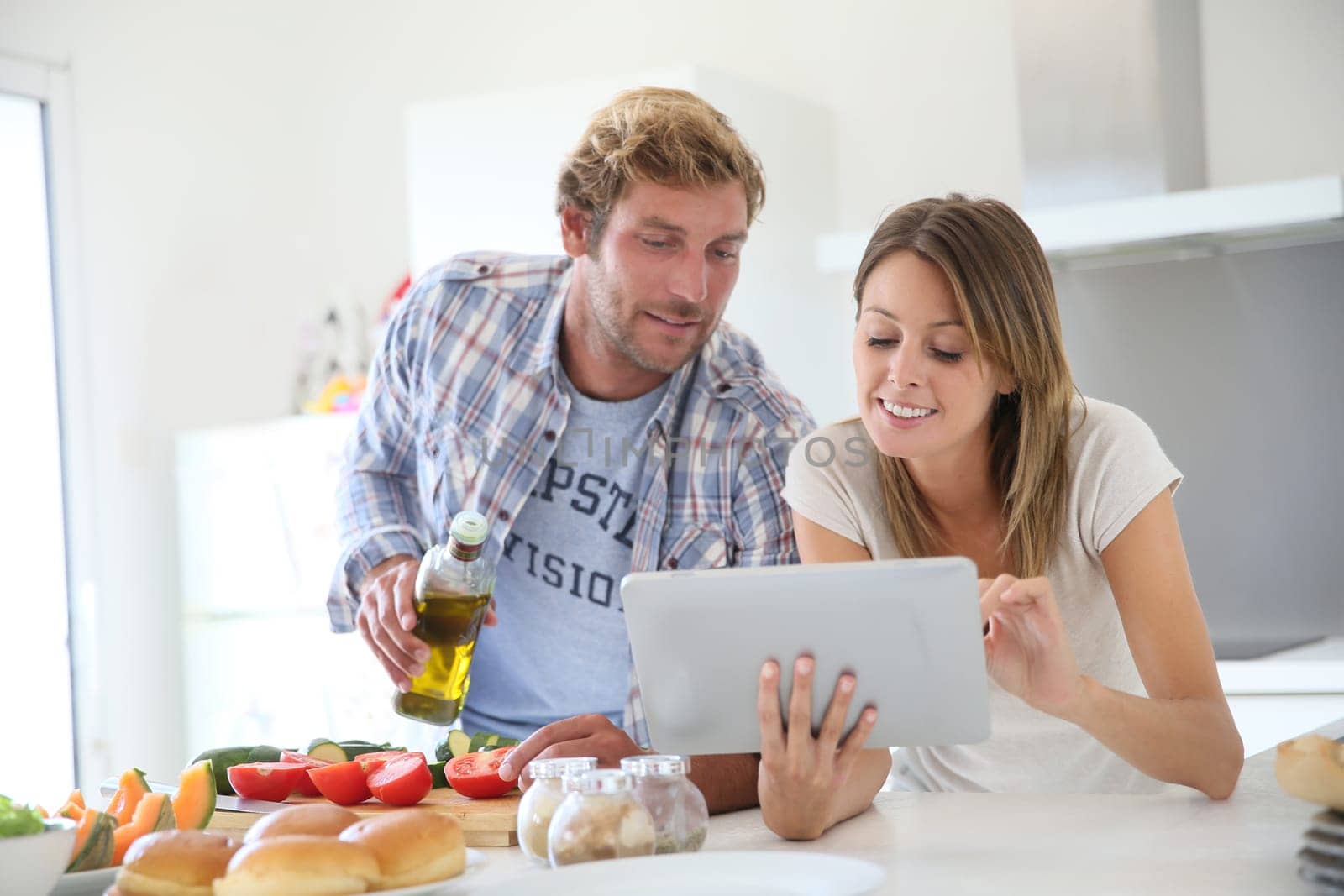 Cheerful couple in kitchen cooking dinner, using tablet