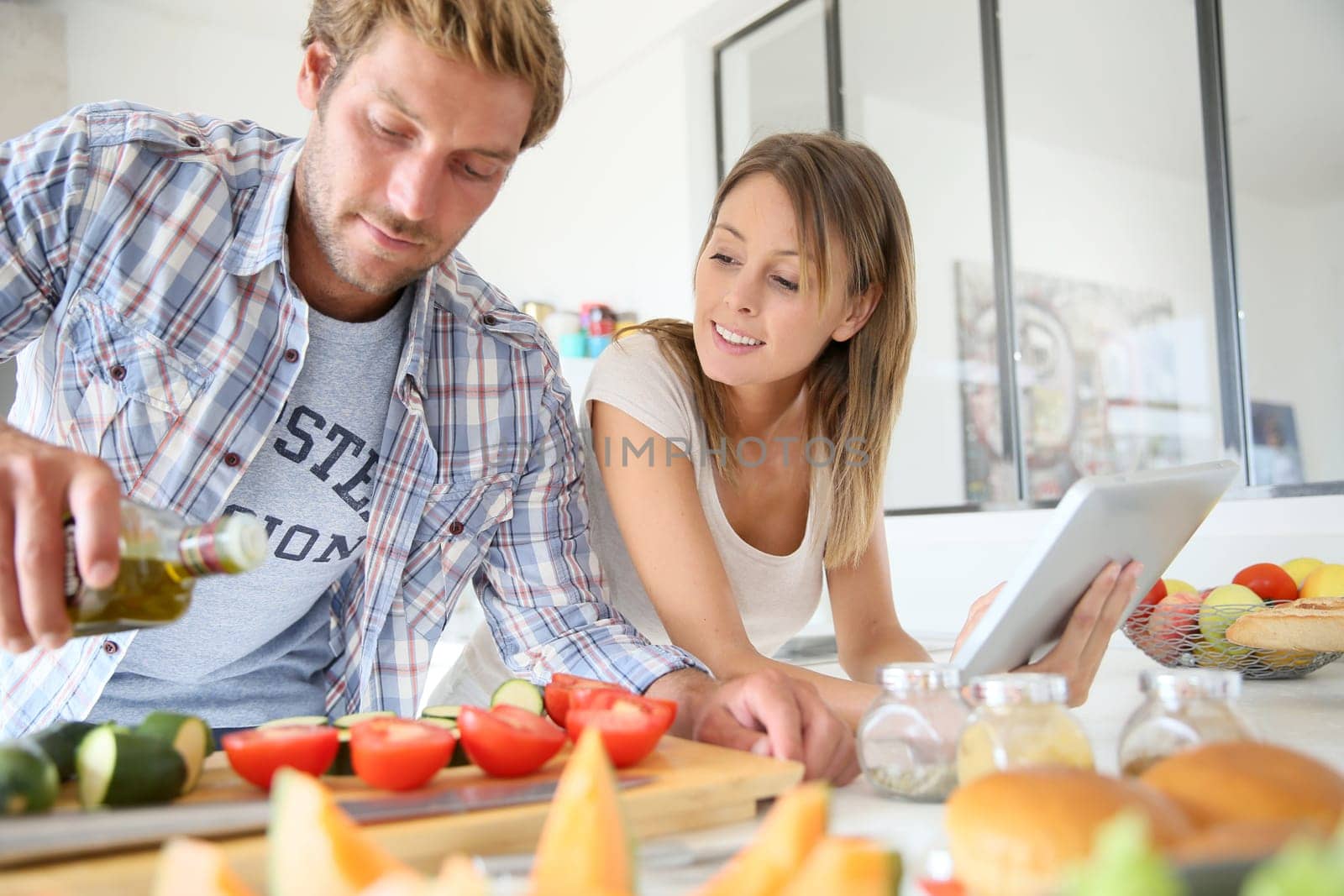 Cheerful couple in kitchen cooking dinner, using tablet