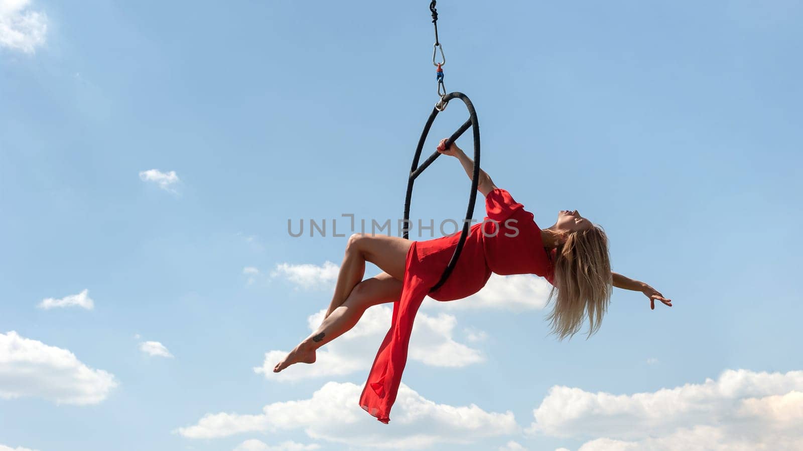 Elegant girl in a red acrobat beautiful dress, performing gymnastic stunts on hoop outdoors against the blue sky with clouds