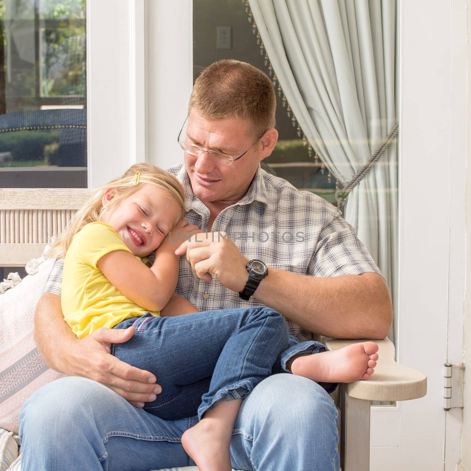 A father and his daughter spend quality time together as they sit on the patio with the father on the bench and his young daughter in his lap.