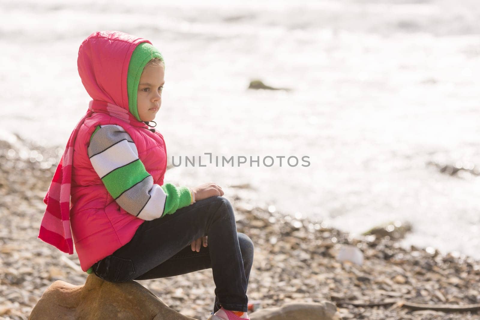 Thoughtful girl sitting on a rock on the stony beach of the sea and looks into the distance